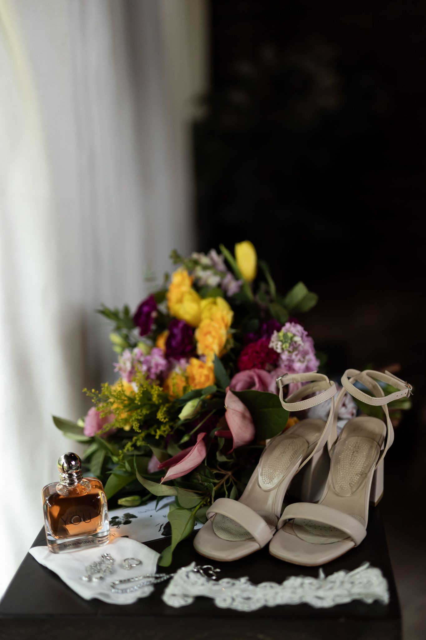 Wedding details featuring bridal bouquet with yellow flowers, white heels, perfume and lace accessories.