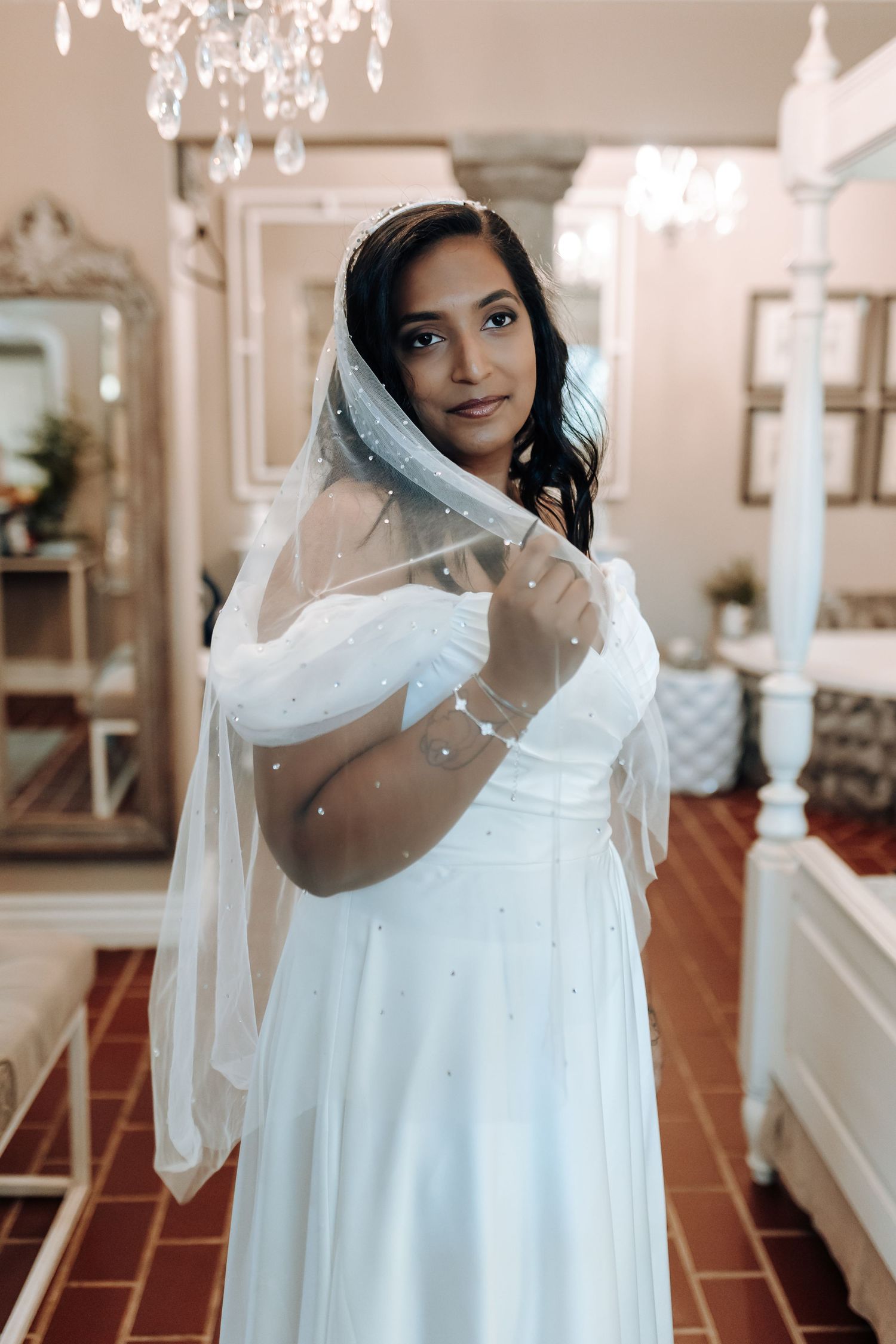 Elegant bride in off-shoulder gown poses with sheer veil in romantic indoor setting.