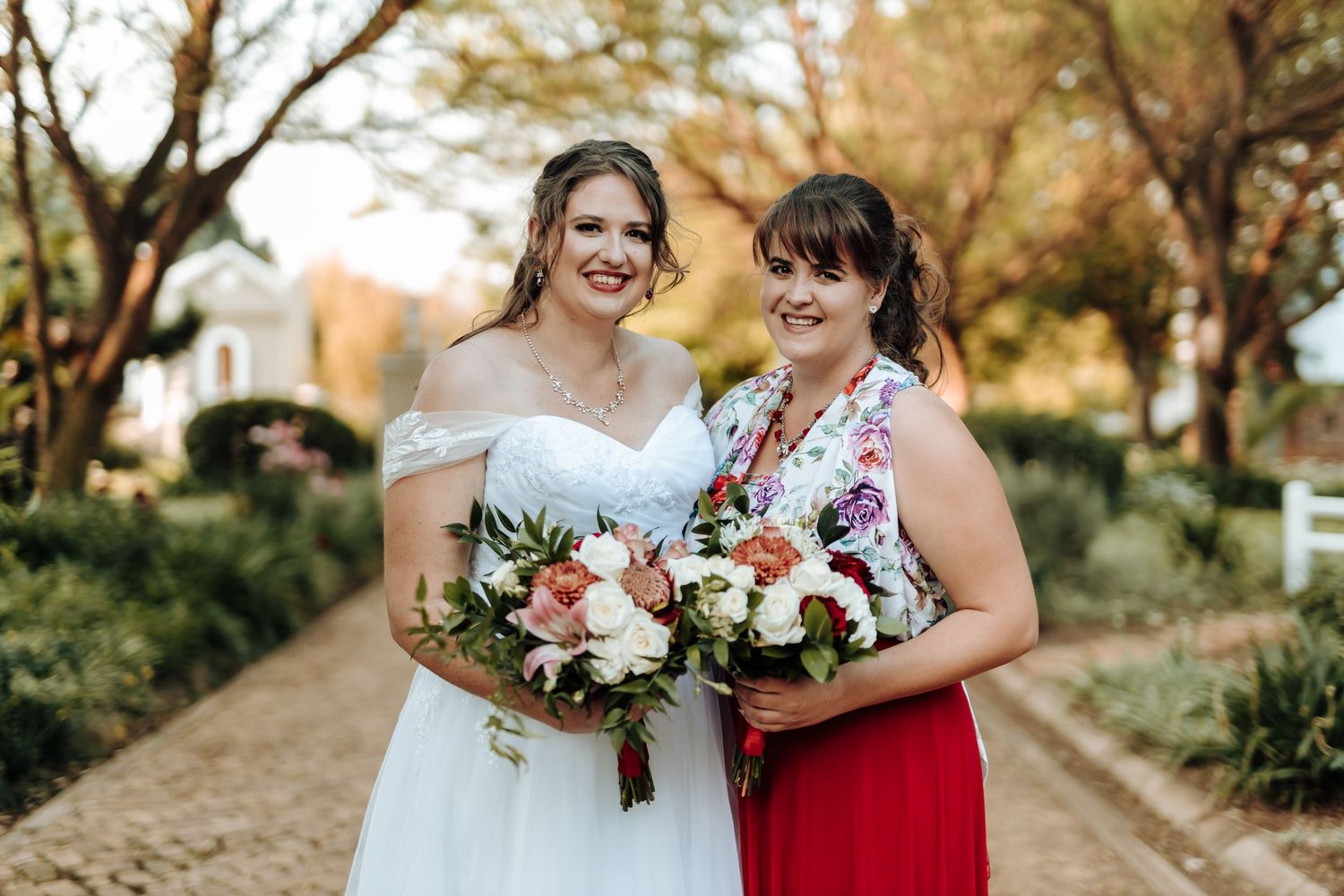 Wedding party members in red and white dresses pose with floral bouquets on brick pathway.