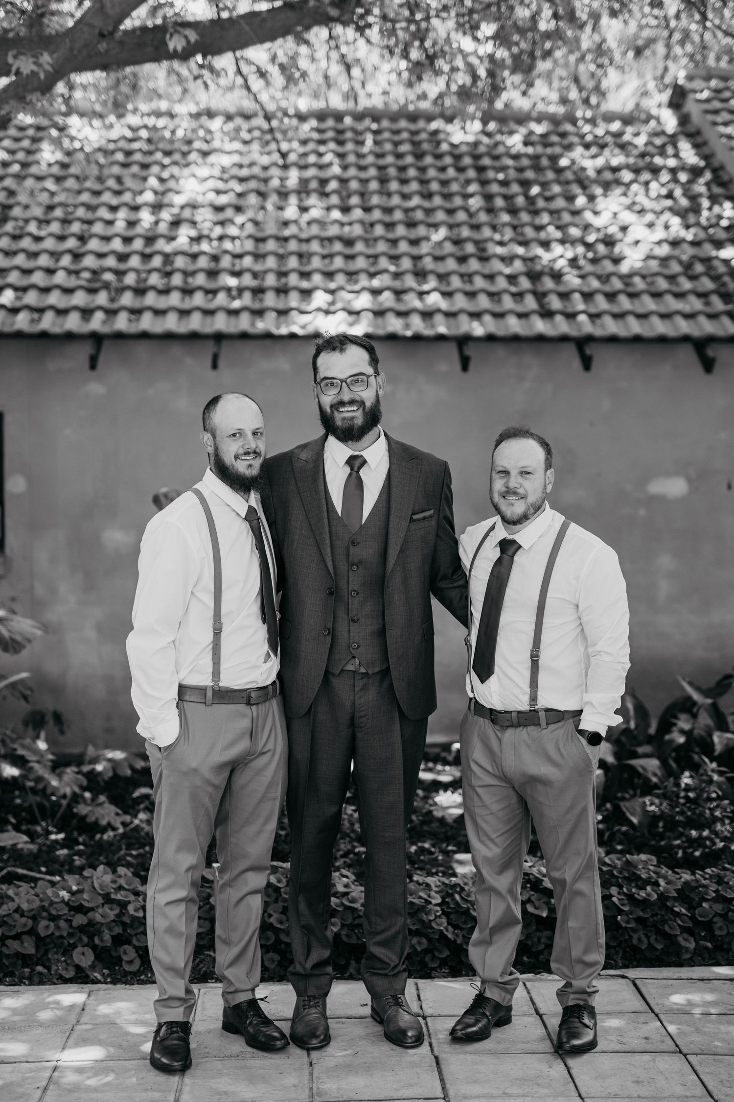 Black and white portrait of three groomsmen in formal attire standing against a building exterior.