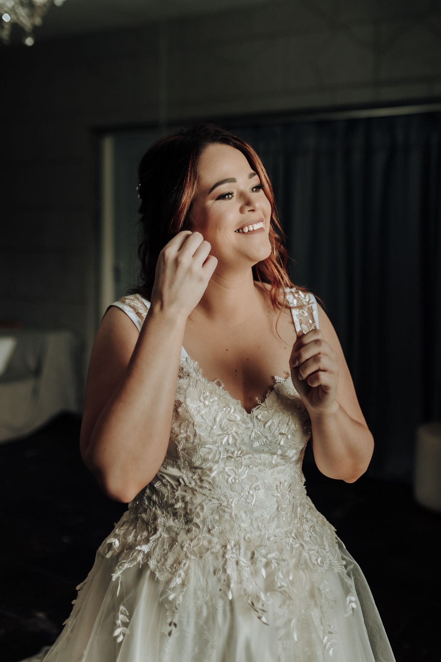 Candid moment of bride in lace wedding dress putting on jewelry with a joyful expression.