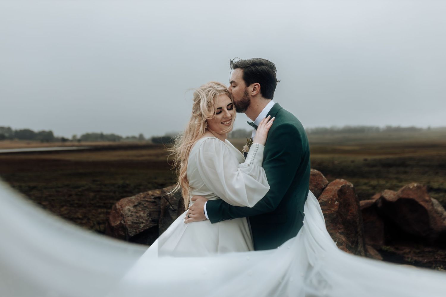 Intimate wedding portrait of couple embracing with flowing veil in dramatic outdoor setting.