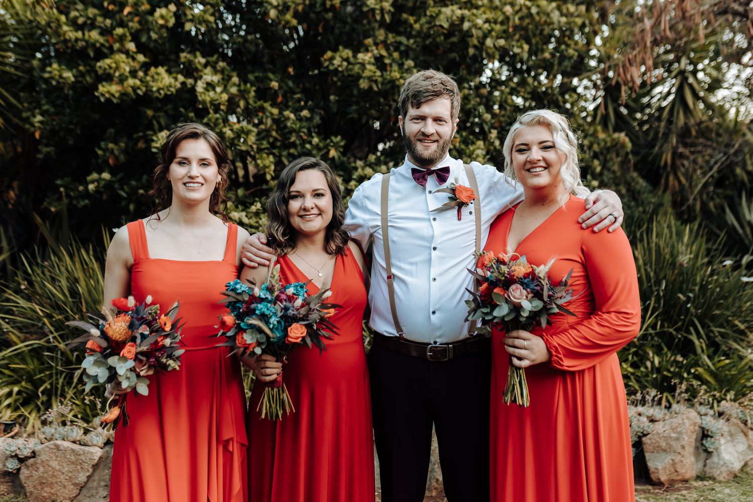 Wedding party members in coral red dresses holding colorful bouquets pose for formal portrait.