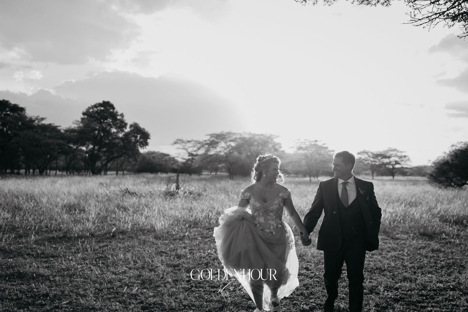 Black and white photo of bride and groom holding hands walking through open field at sunset.