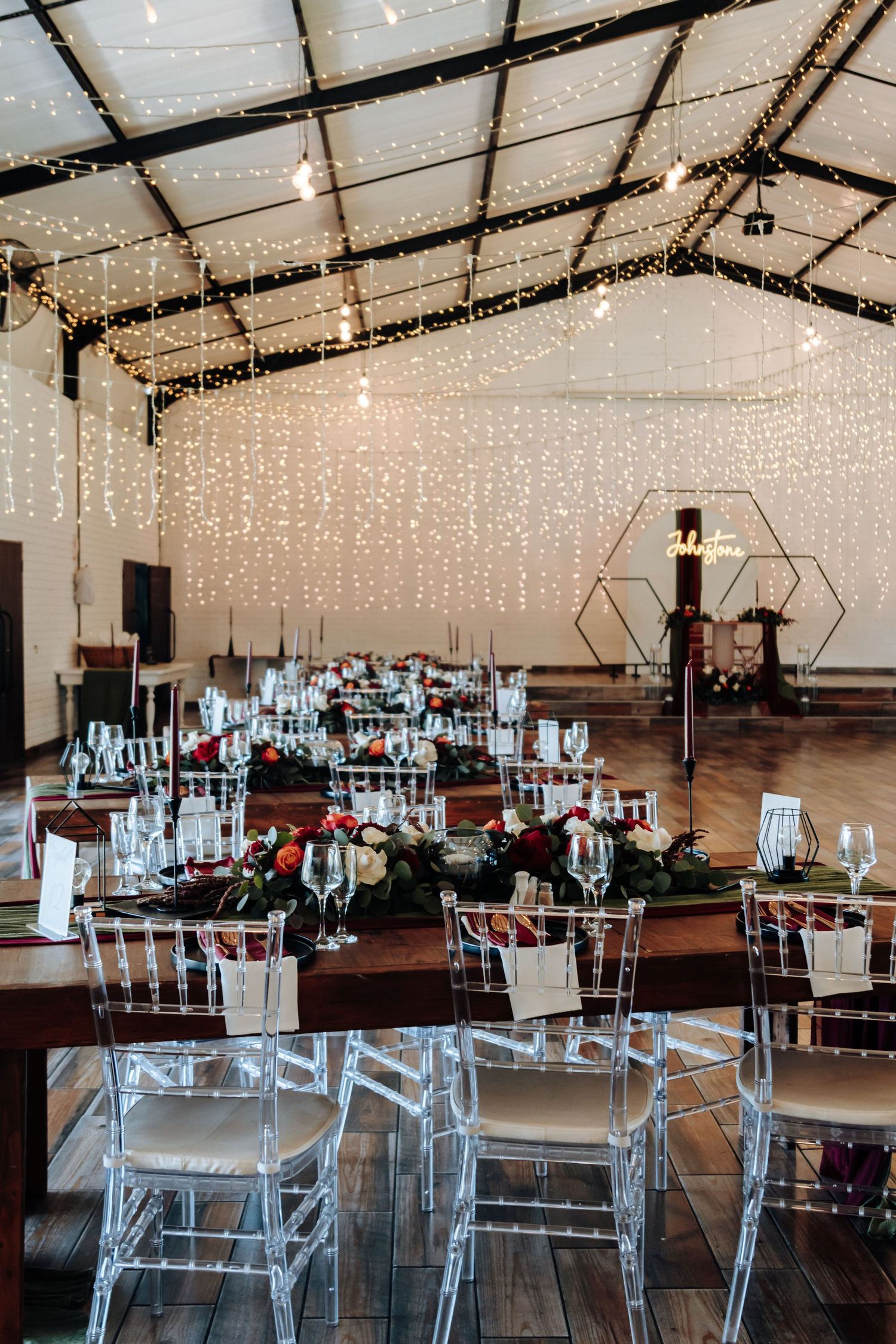 Elegant wedding reception venue with fairy lights on ceiling, crystal chairs, floral centerpieces, and geometric decor on wood floors.