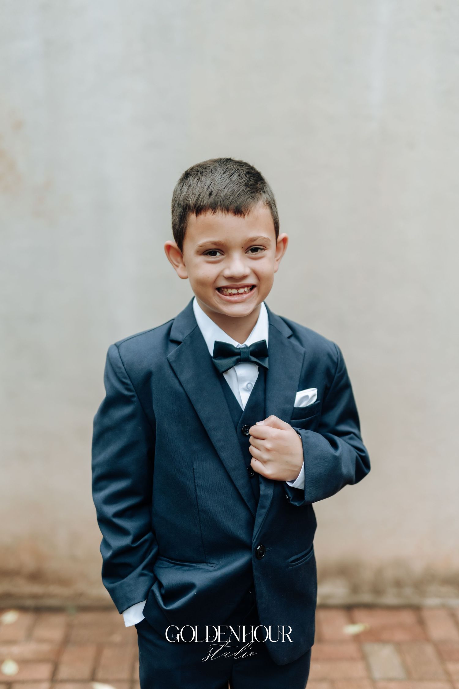 Adorable young boy in a navy blue suit and bow tie smiling at a wedding ceremony.