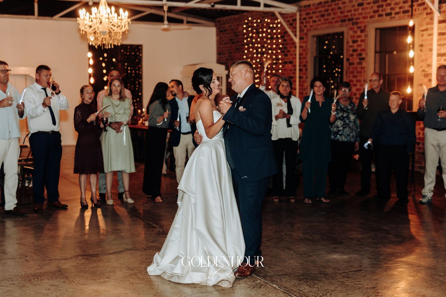 Bride and groom share first dance in rustic venue with chandelier and string lights, guests watching.