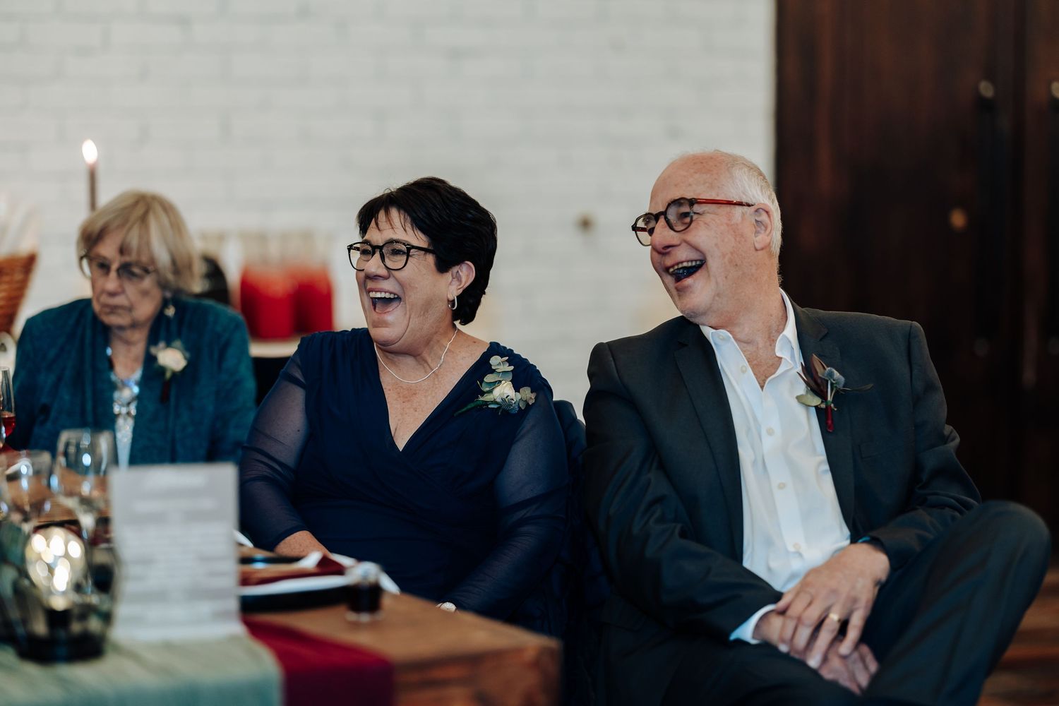 Two guests laughing joyfully at a wedding reception, seated at a decorated table in an elegant venue.