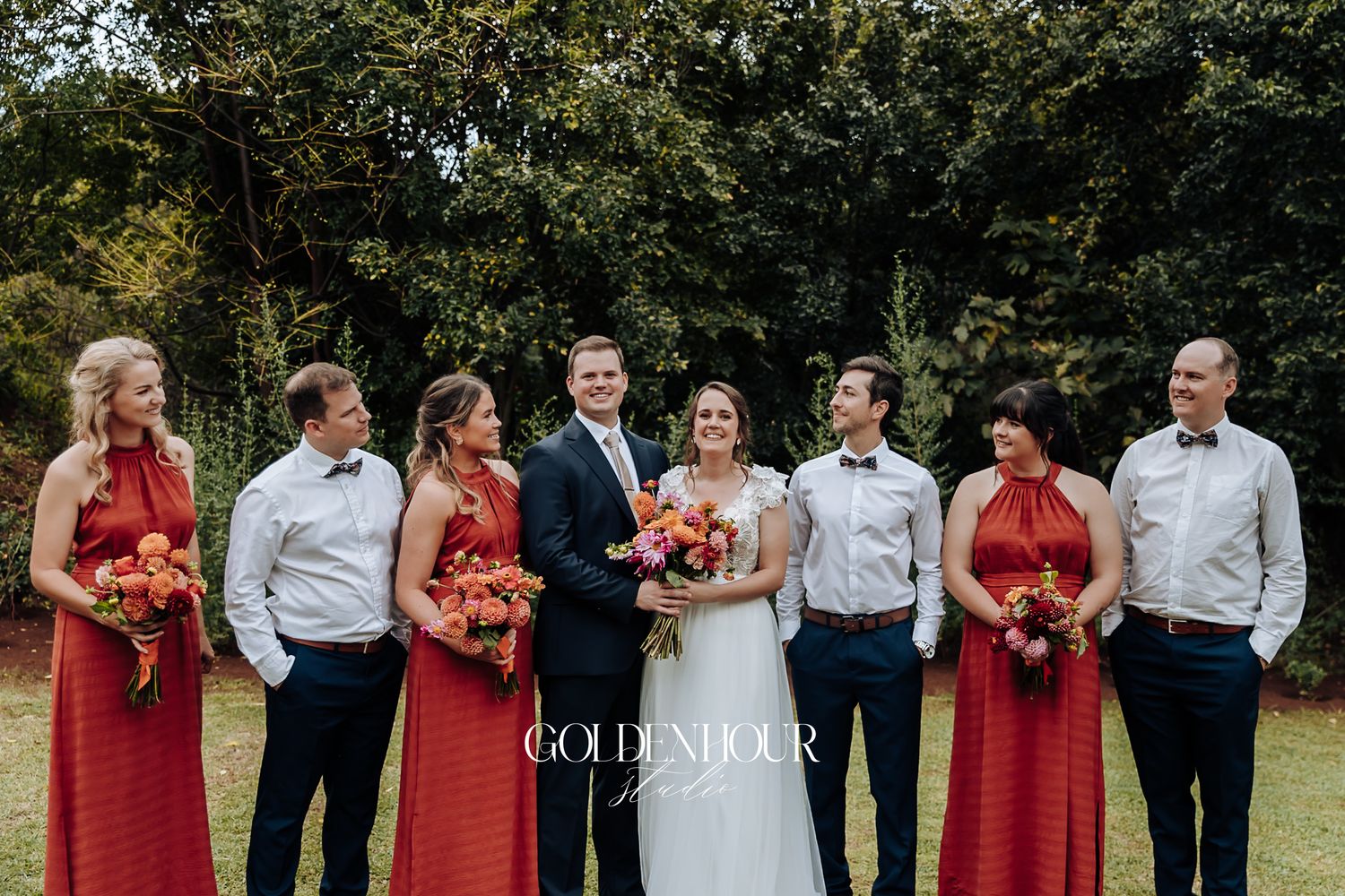 Wedding party in red dresses and navy pants laughing together on lush green lawn outdoors.