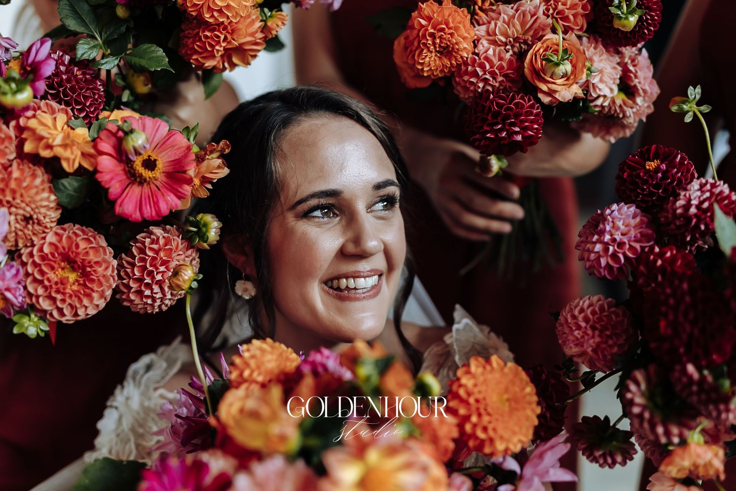 Smiling bride surrounded by lush orange, pink, and red dahlia wedding bouquets in a close-up portrait.