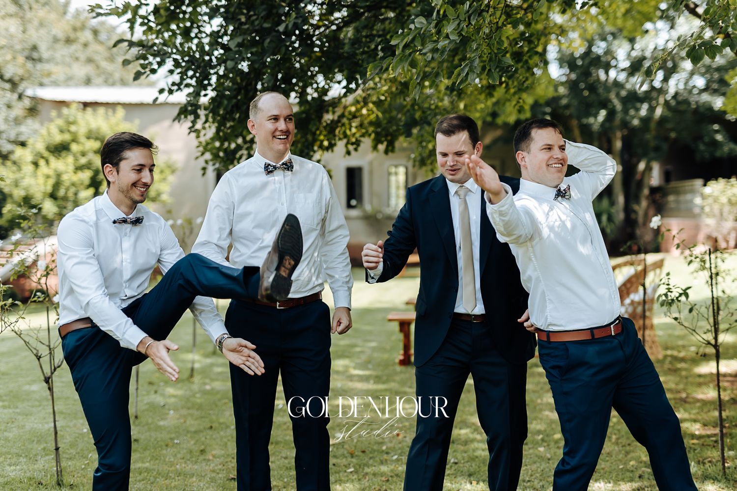 Groomsmen laughing and posing playfully outdoors under a tree at a wedding.