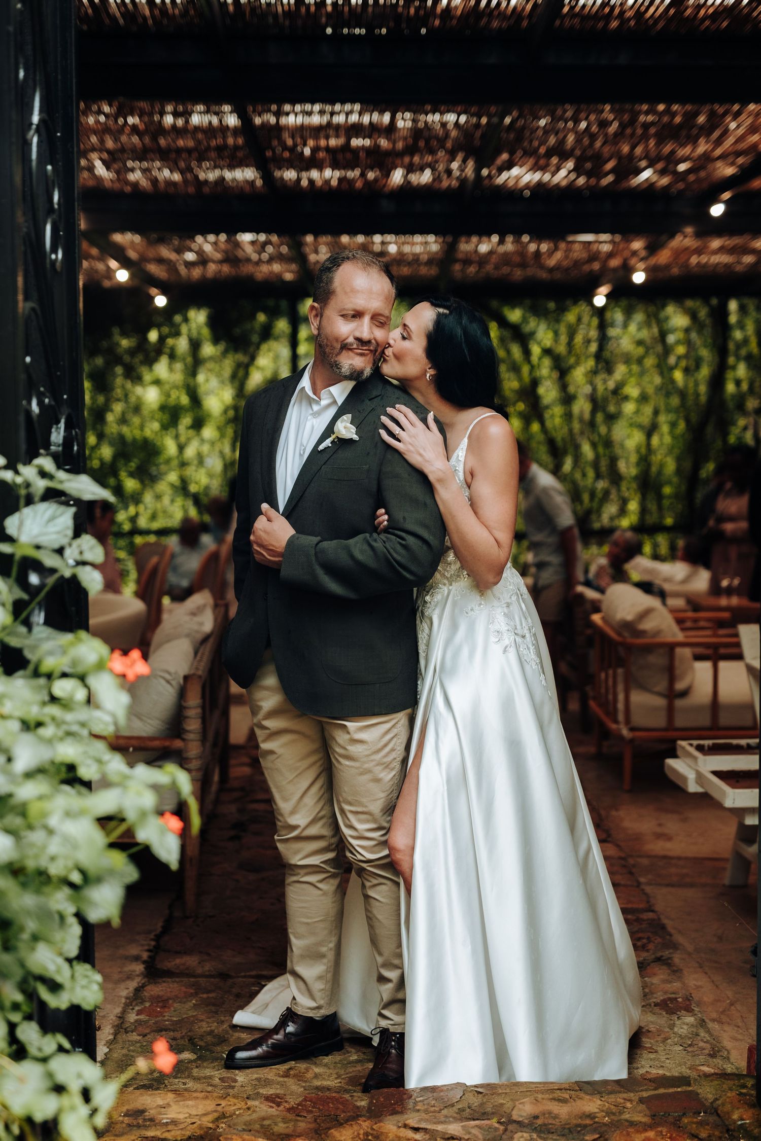 Bride in white gown embraces groom in dark blazer at outdoor wedding venue with lush greenery.