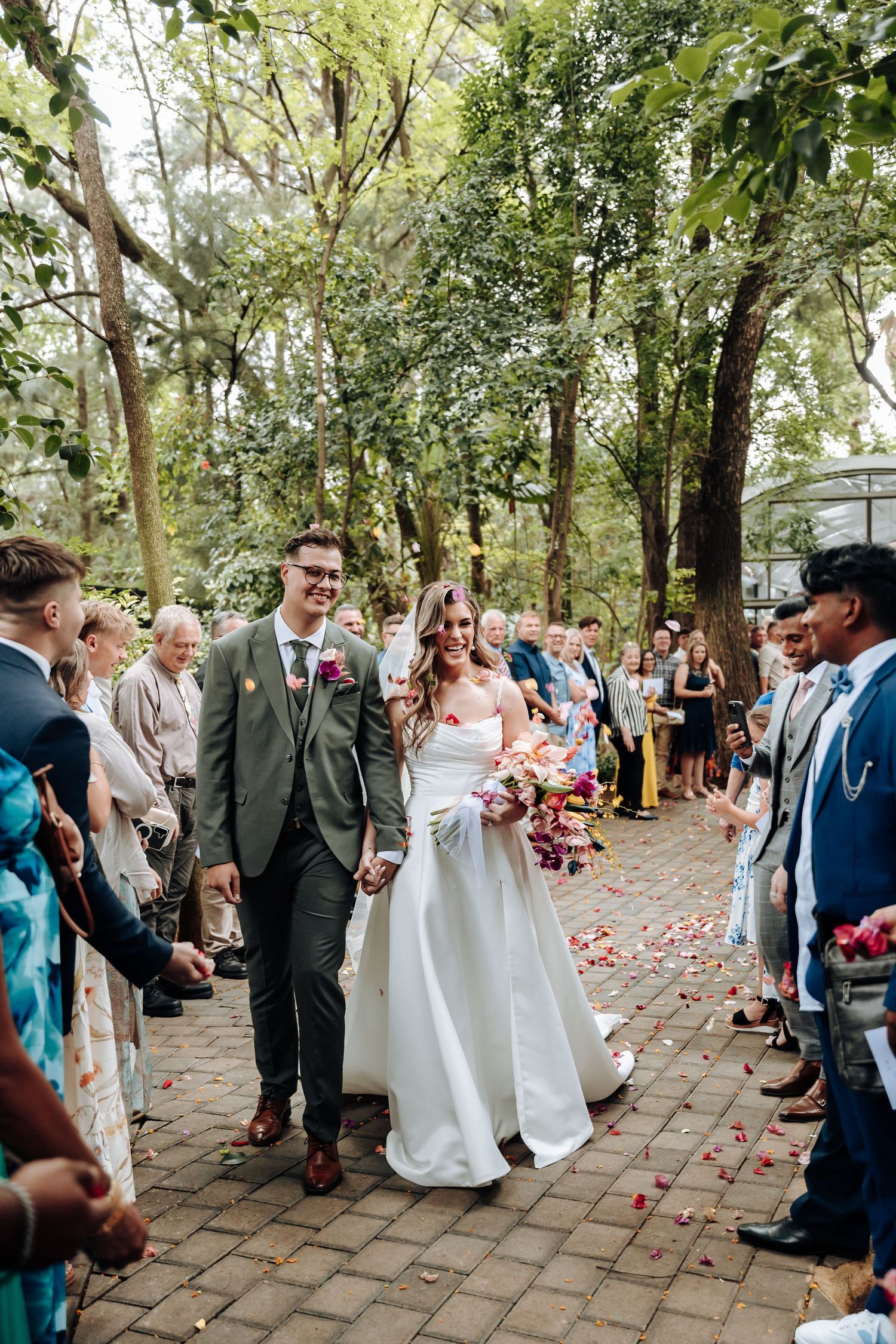 Wedding ceremony on a brick pathway surrounded by lush green trees with rose petals scattered on the ground.