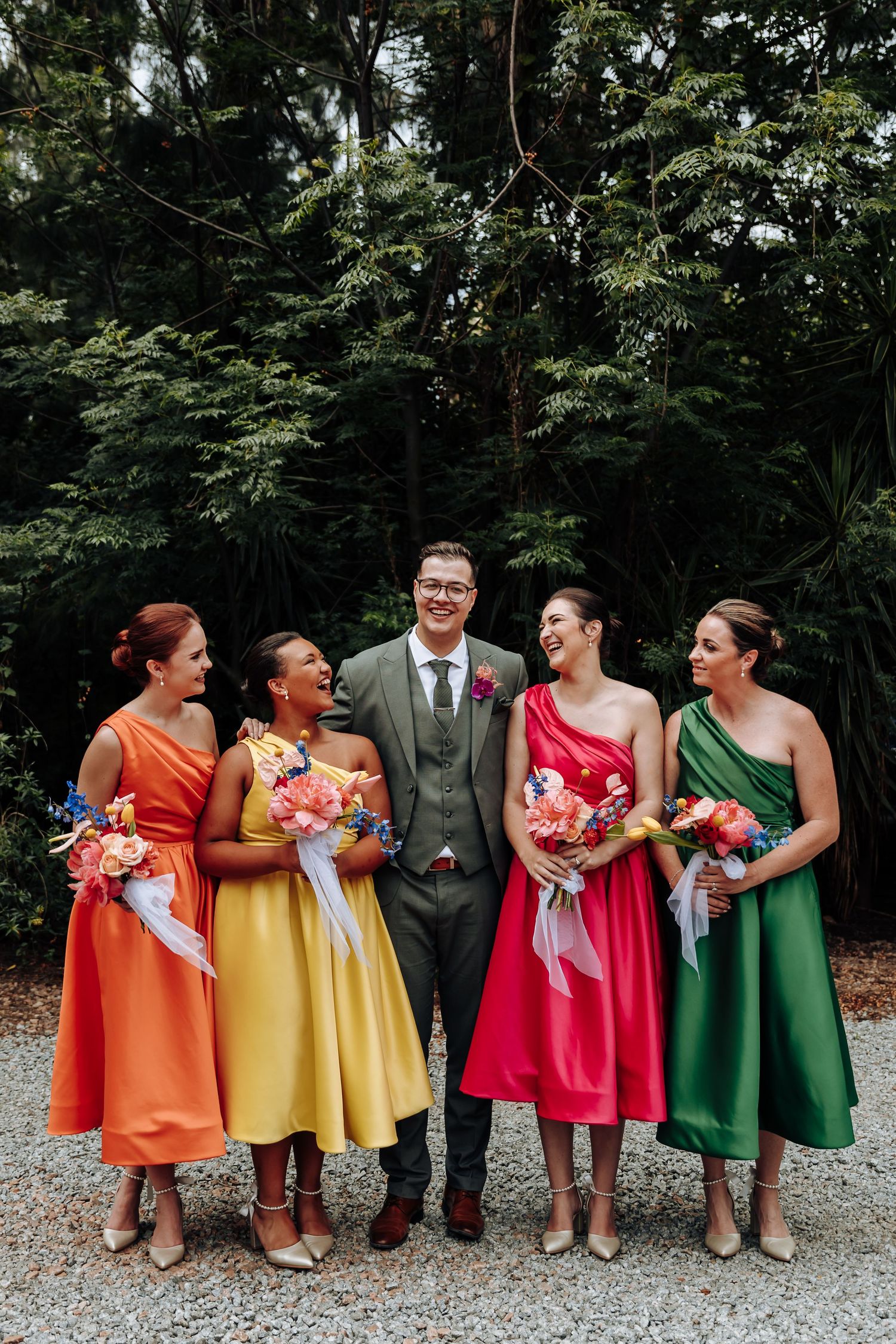 Wedding party wearing colorful rainbow dresses in orange, yellow, red and green stand together outdoors.