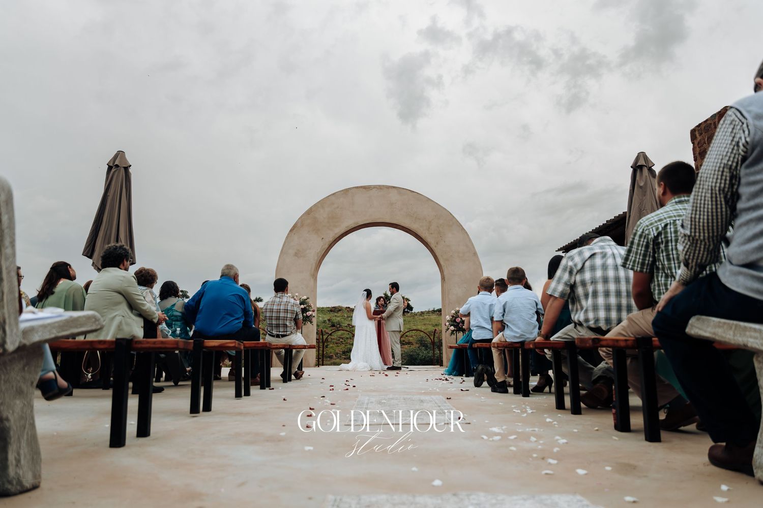 Panoramic view of guests gathered at an outdoor wedding ceremony venue with stone arches against a cloudy sky.