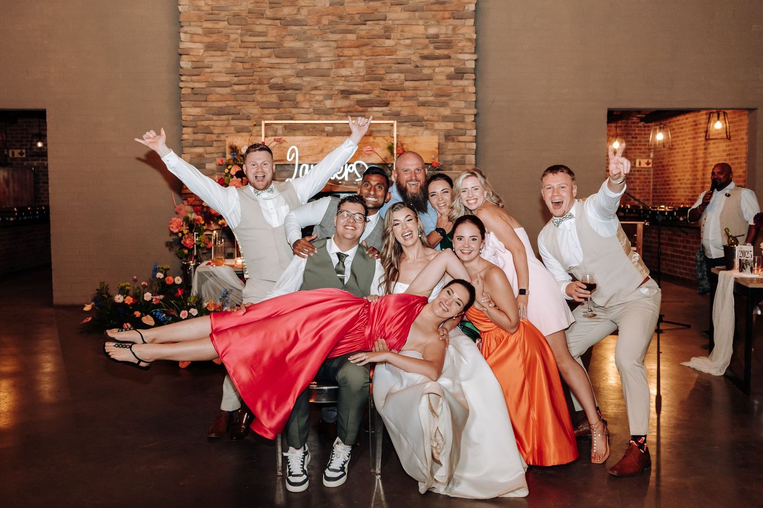 Wedding group strikes playful poses for a photo with colorful dresses against stone wall.