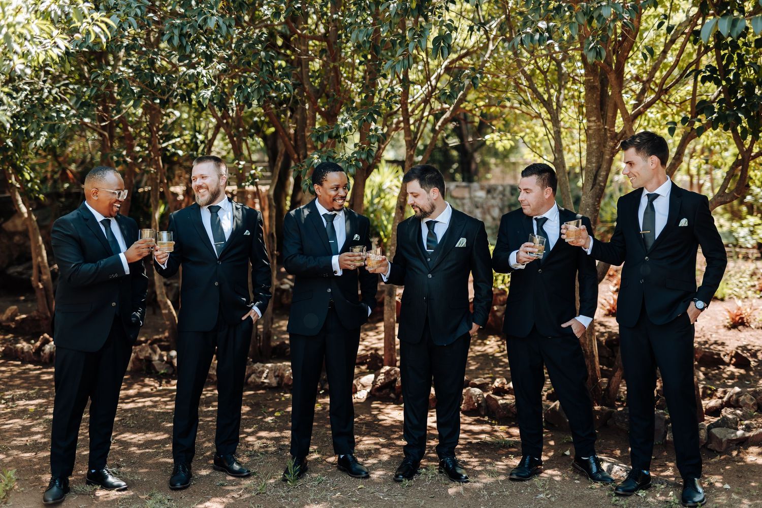 Groomsmen in black suits pose together outdoors in a wooded setting during a wedding celebration.