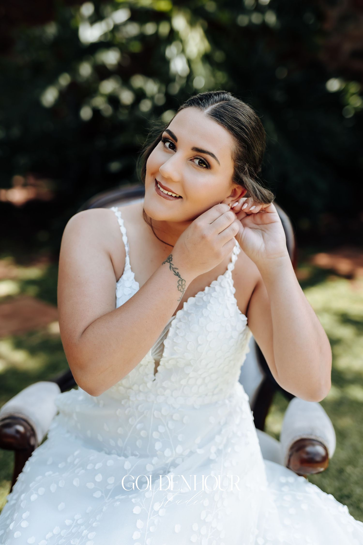 A bride wearing a white lace wedding dress sits outdoors putting on earrings.