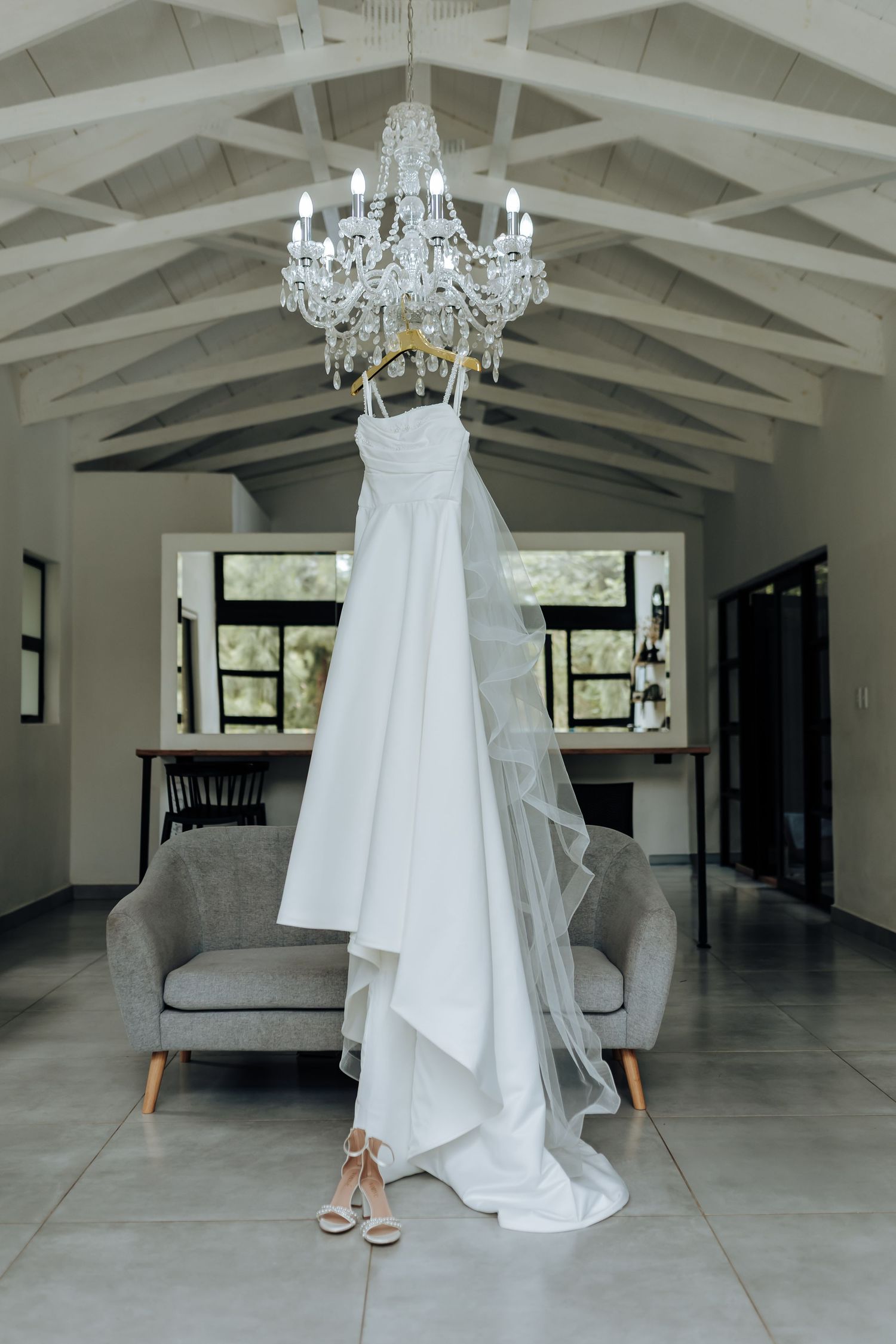 White wedding dress hangs elegantly beneath a crystal chandelier in a bright modern space.