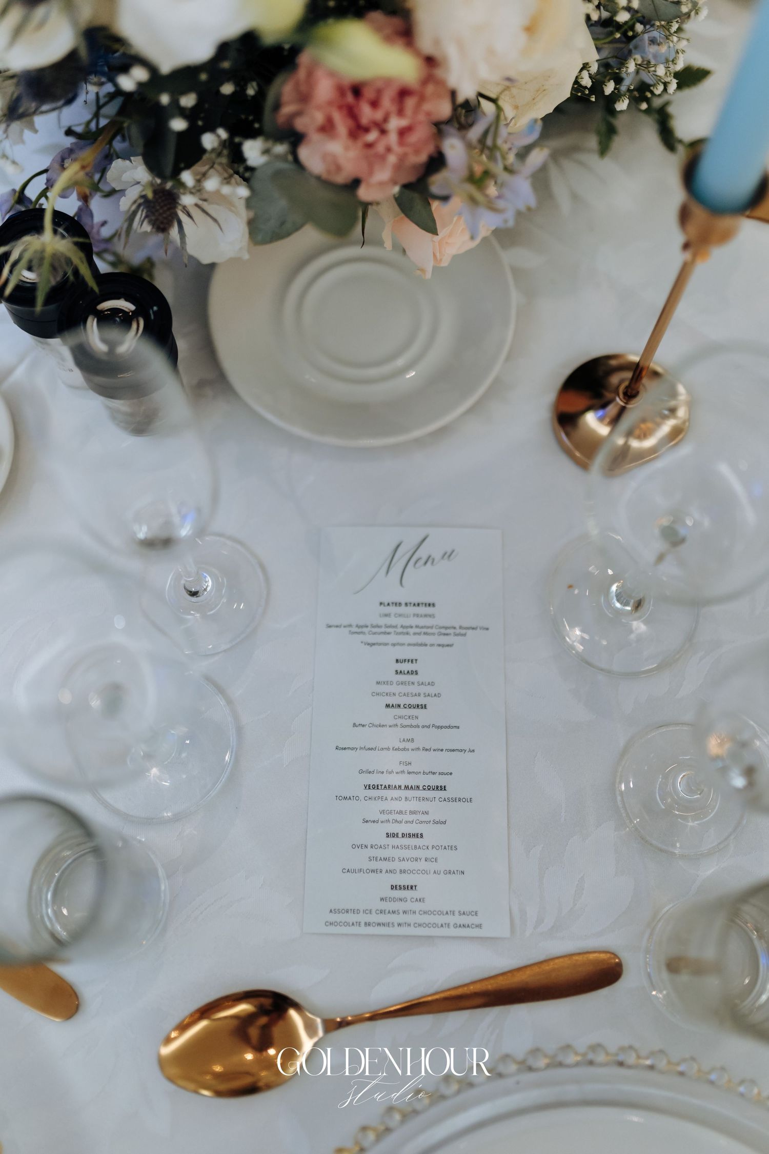 Wedding menu card surrounded by wine glasses and floral arrangements on white tablecloth.