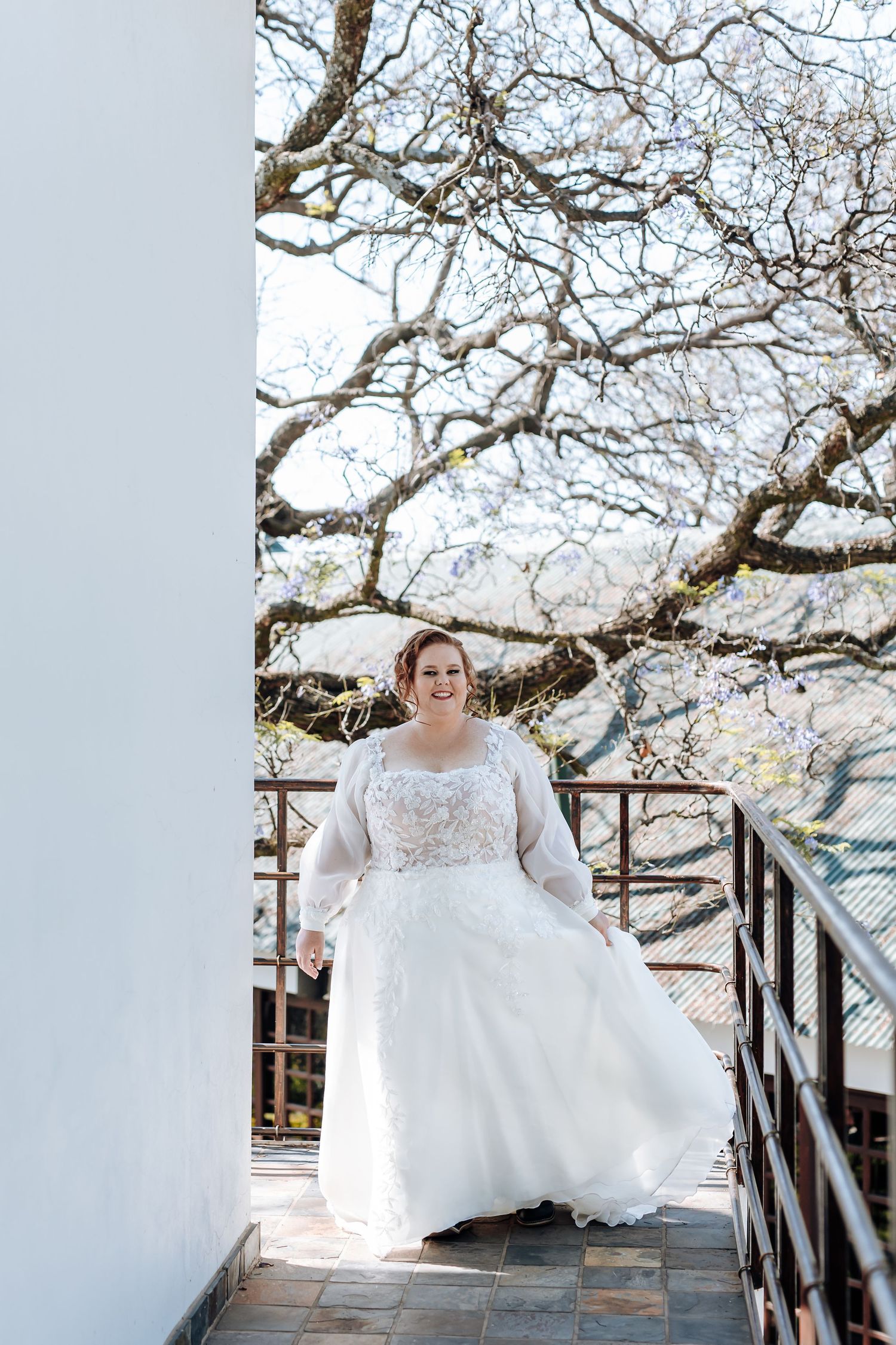 A bride in a white lace wedding dress poses on a wooden deck surrounded by blooming cherry blossom trees.