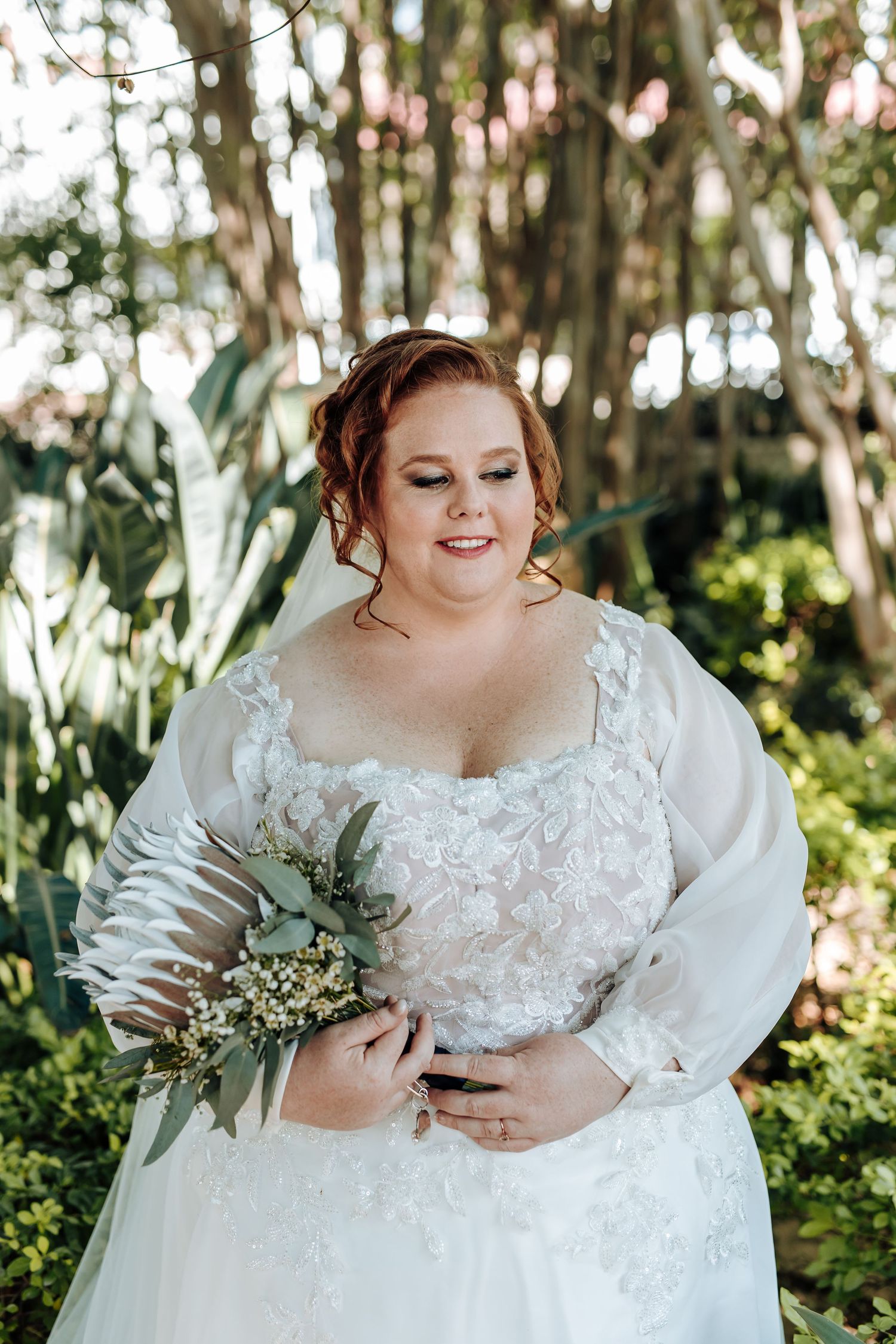 A bride in a detailed lace wedding gown holds a delicate bouquet of white flowers and greenery in a sunlit forest setting.