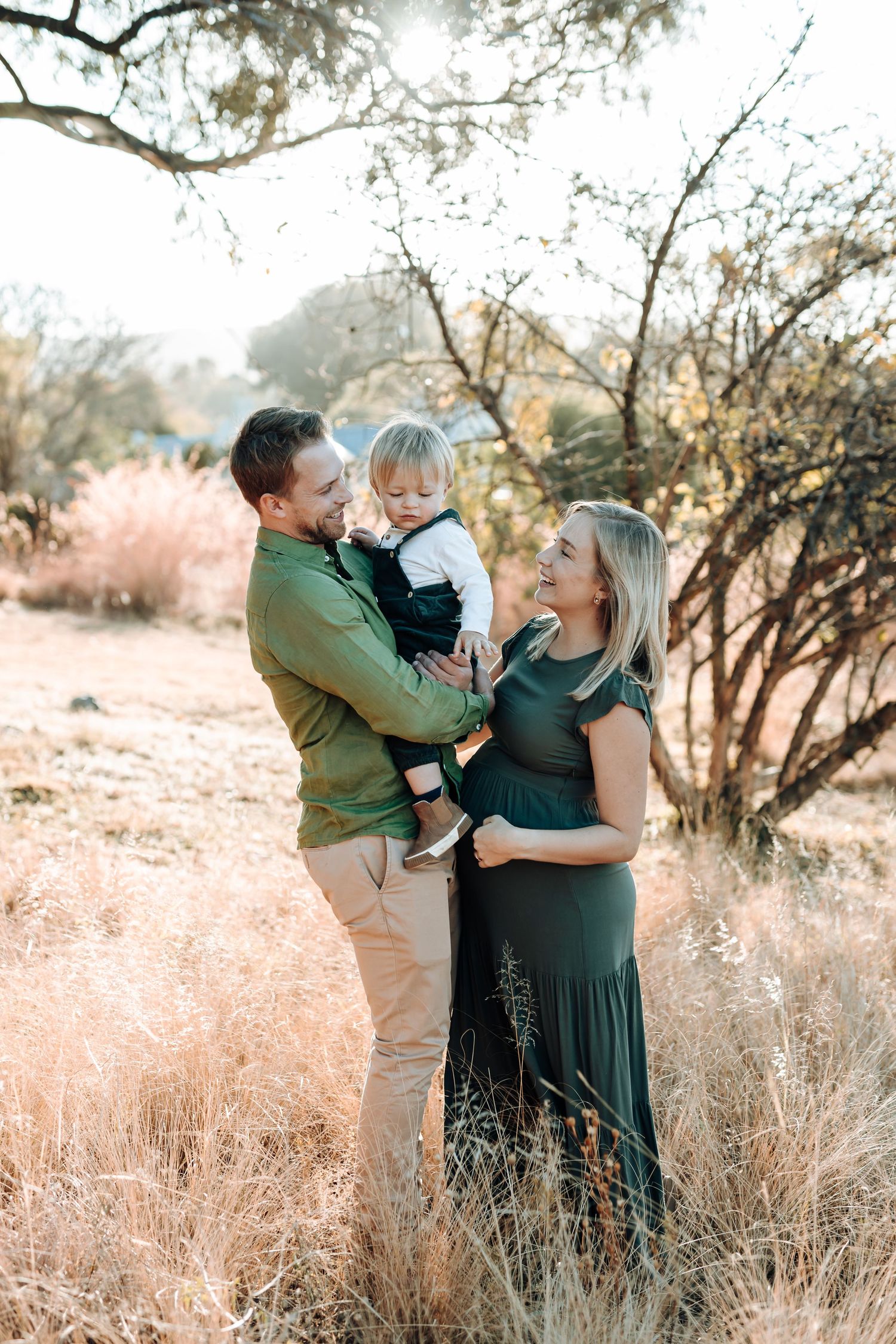 Family shares tender moments together in a sun-drenched field with golden grass and trees at sunset.