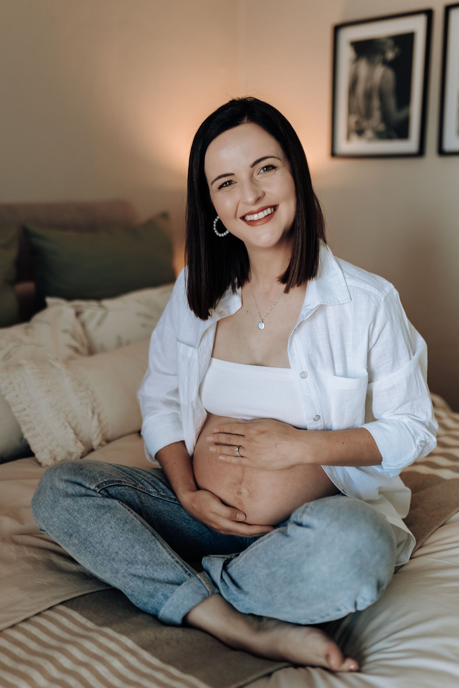 Expectant mother in white shirt relaxes on bed with gentle belly touches in warm light setting.