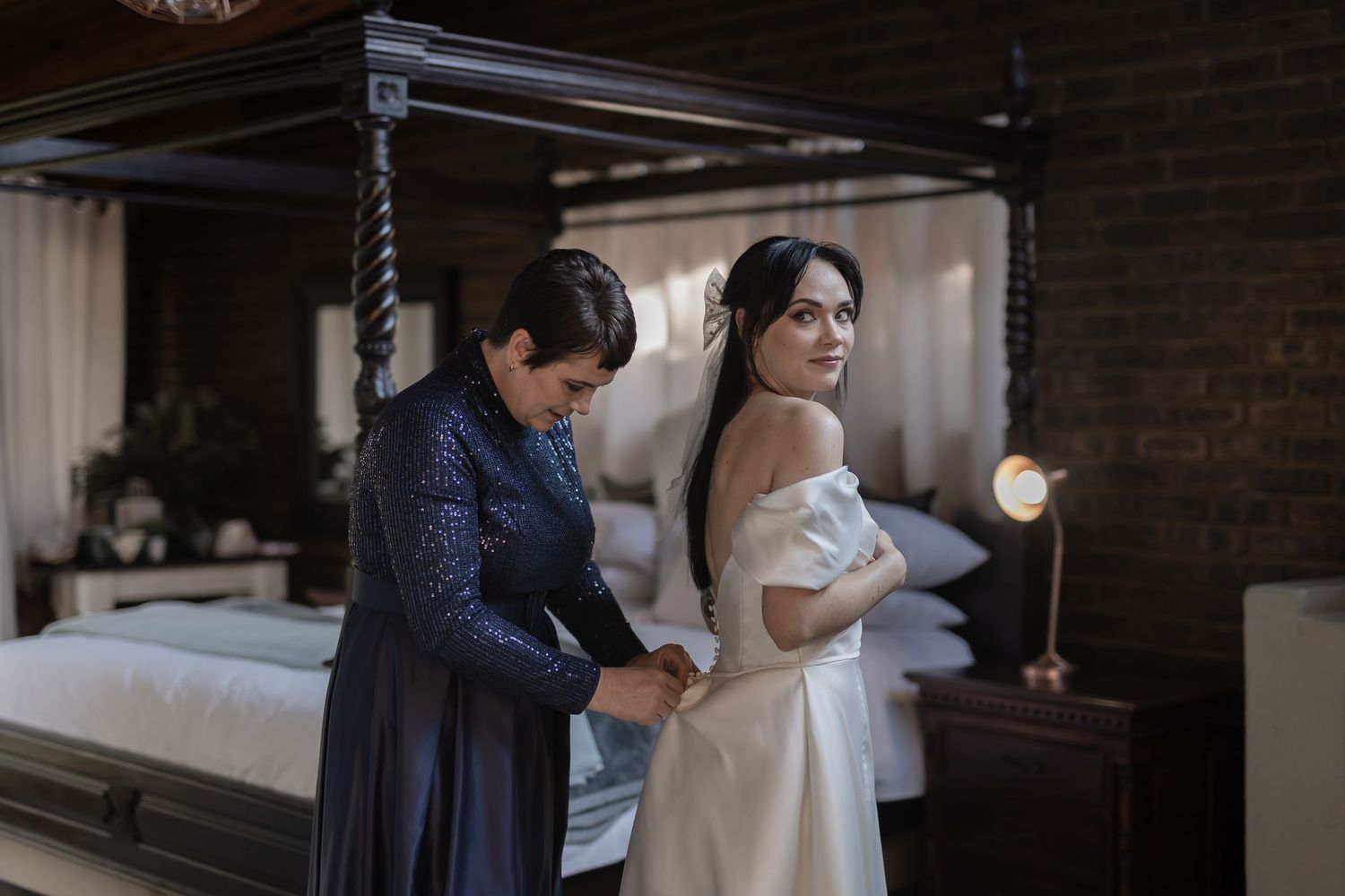 Two figures stand in an intimate moment during wedding preparations in a rustic bedroom setting.