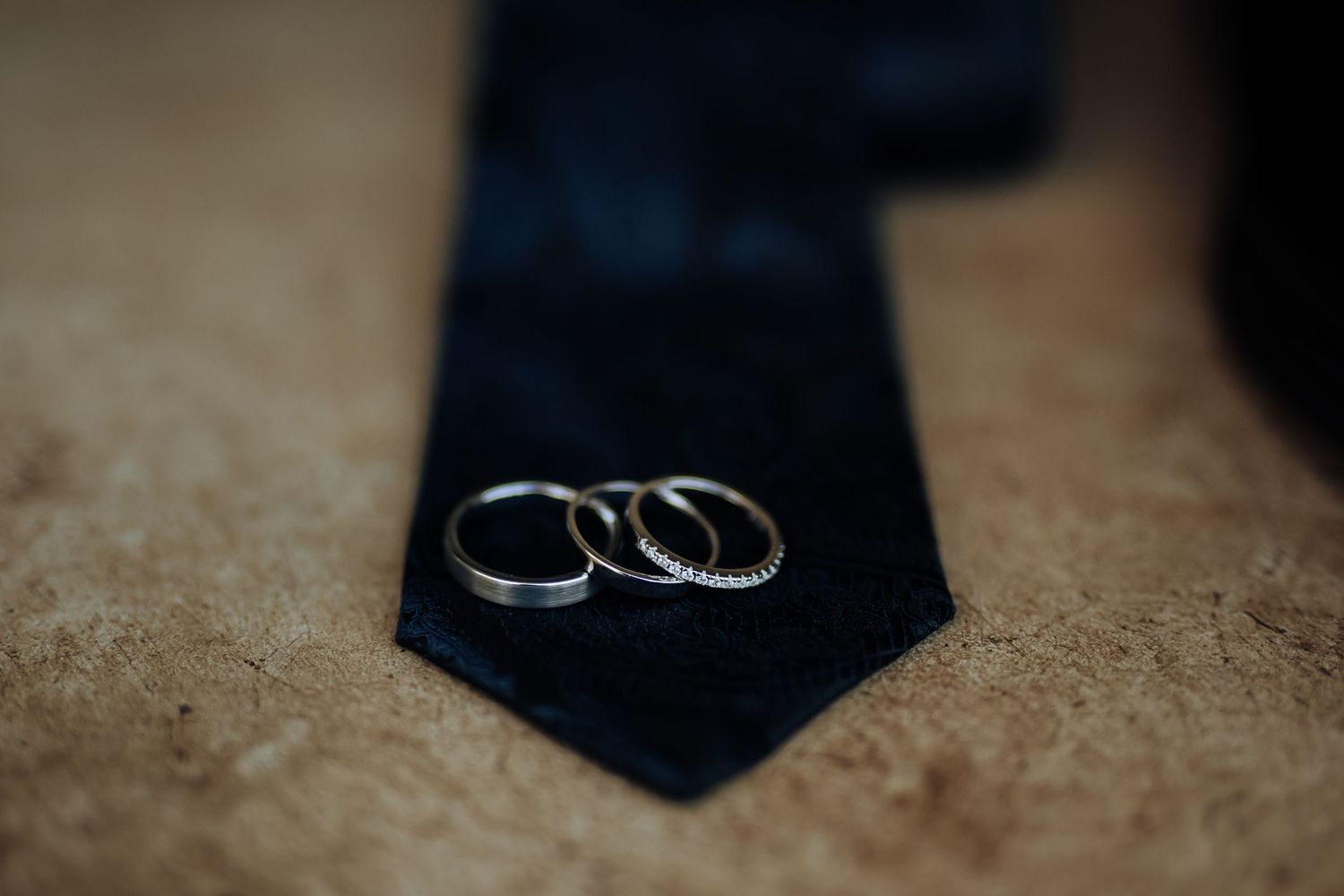 Two wedding rings rest on a black necktie against a textured beige background in a macro shot.