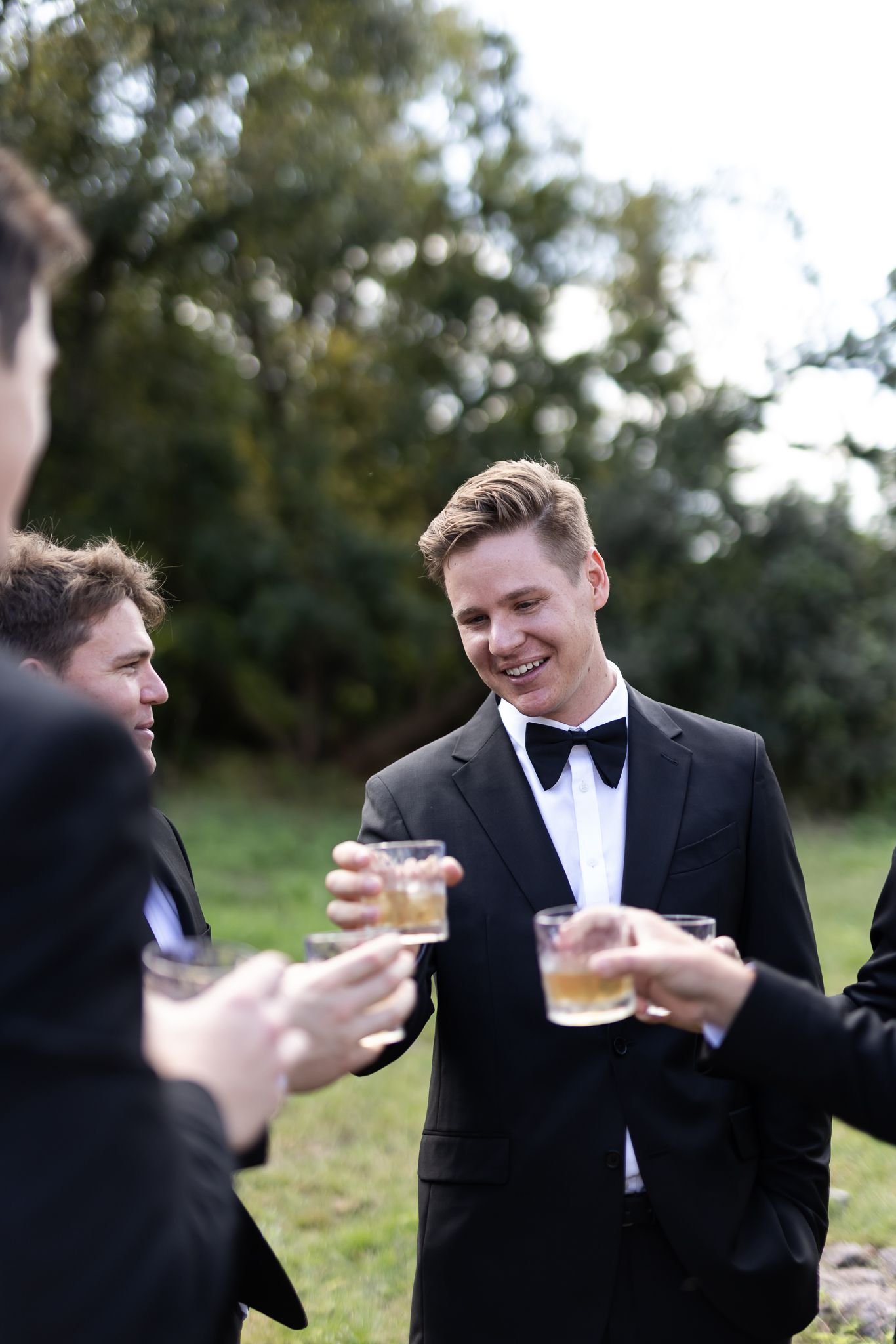 Groomsmen in tuxedos sharing drinks and laughing at outdoor event.