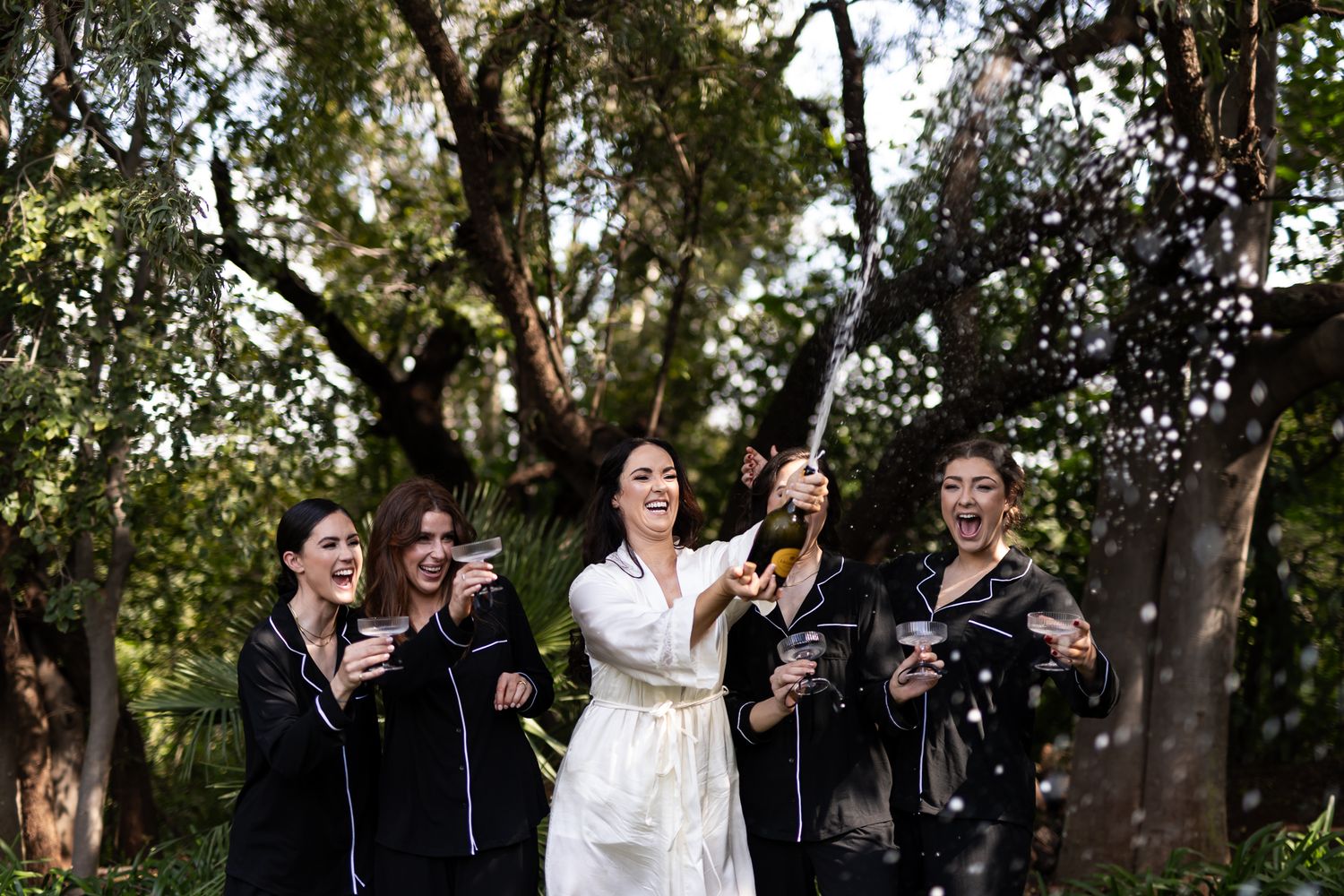 Bride in white dress celebrates with bridesmaids in black robes holding champagne glasses in outdoor garden setting.