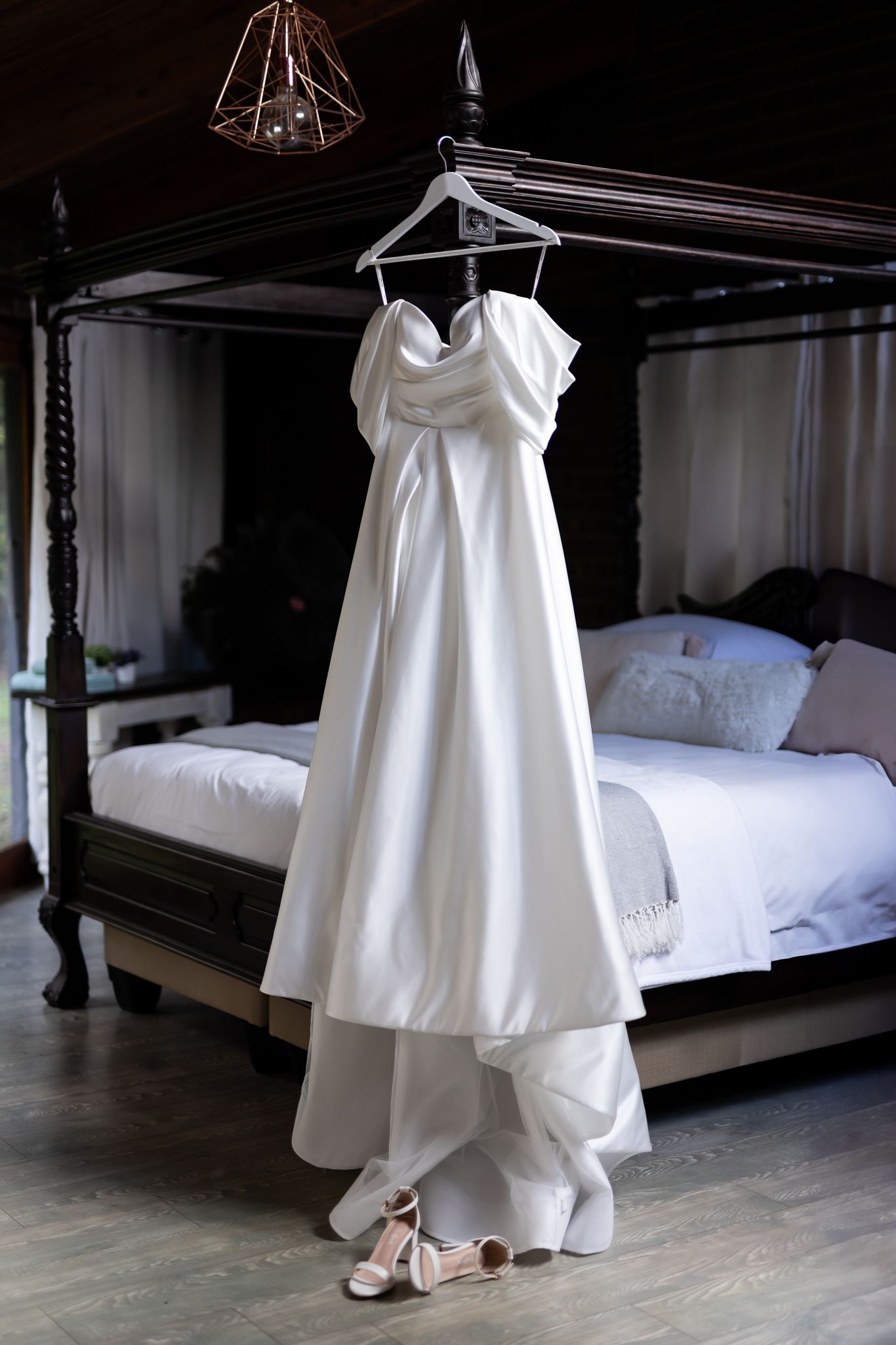 Elegant white wedding dress hanging on four-poster bed frame in dimly lit bedroom.