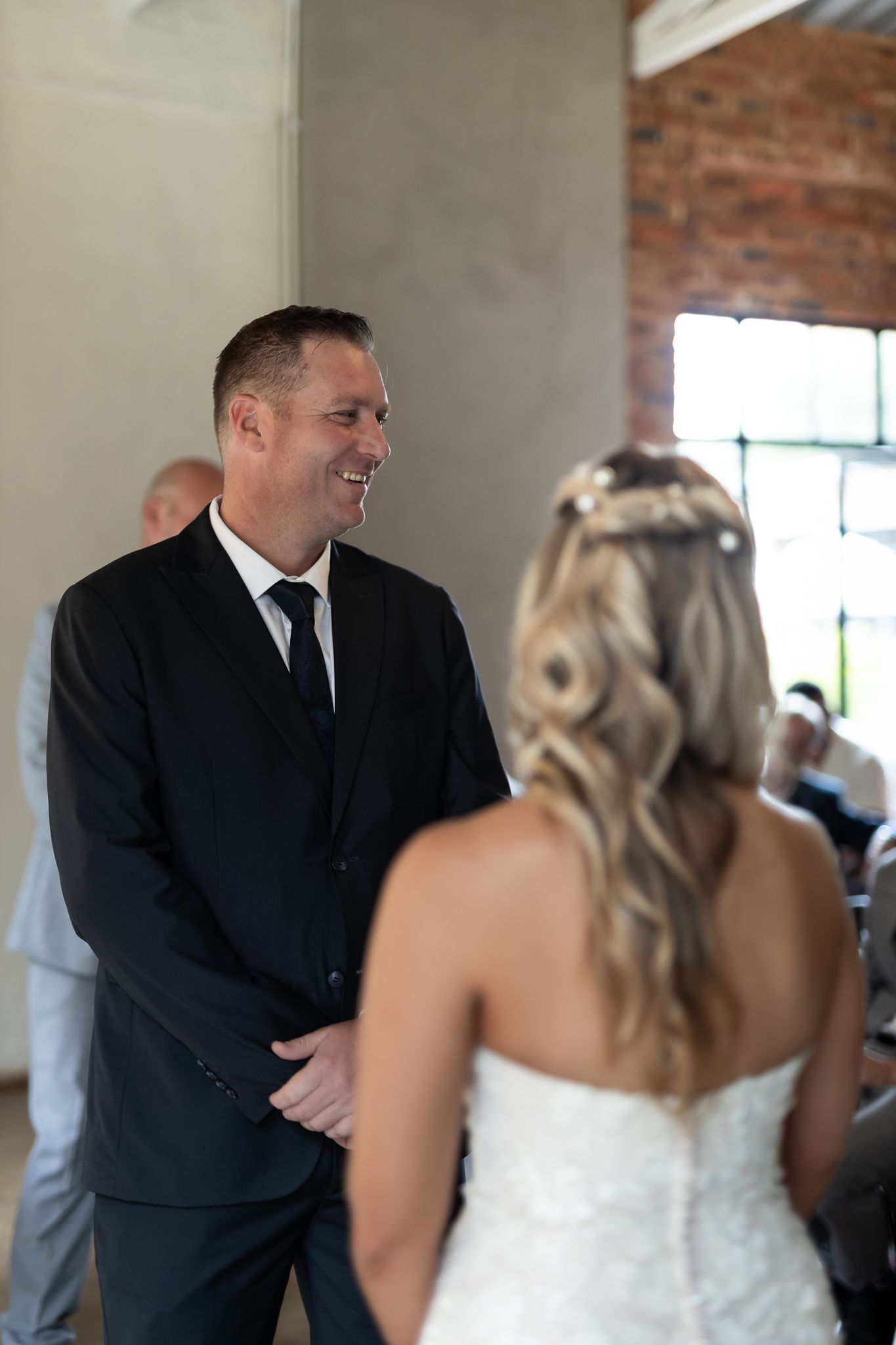 A groom in a black suit smiles during the wedding ceremony.