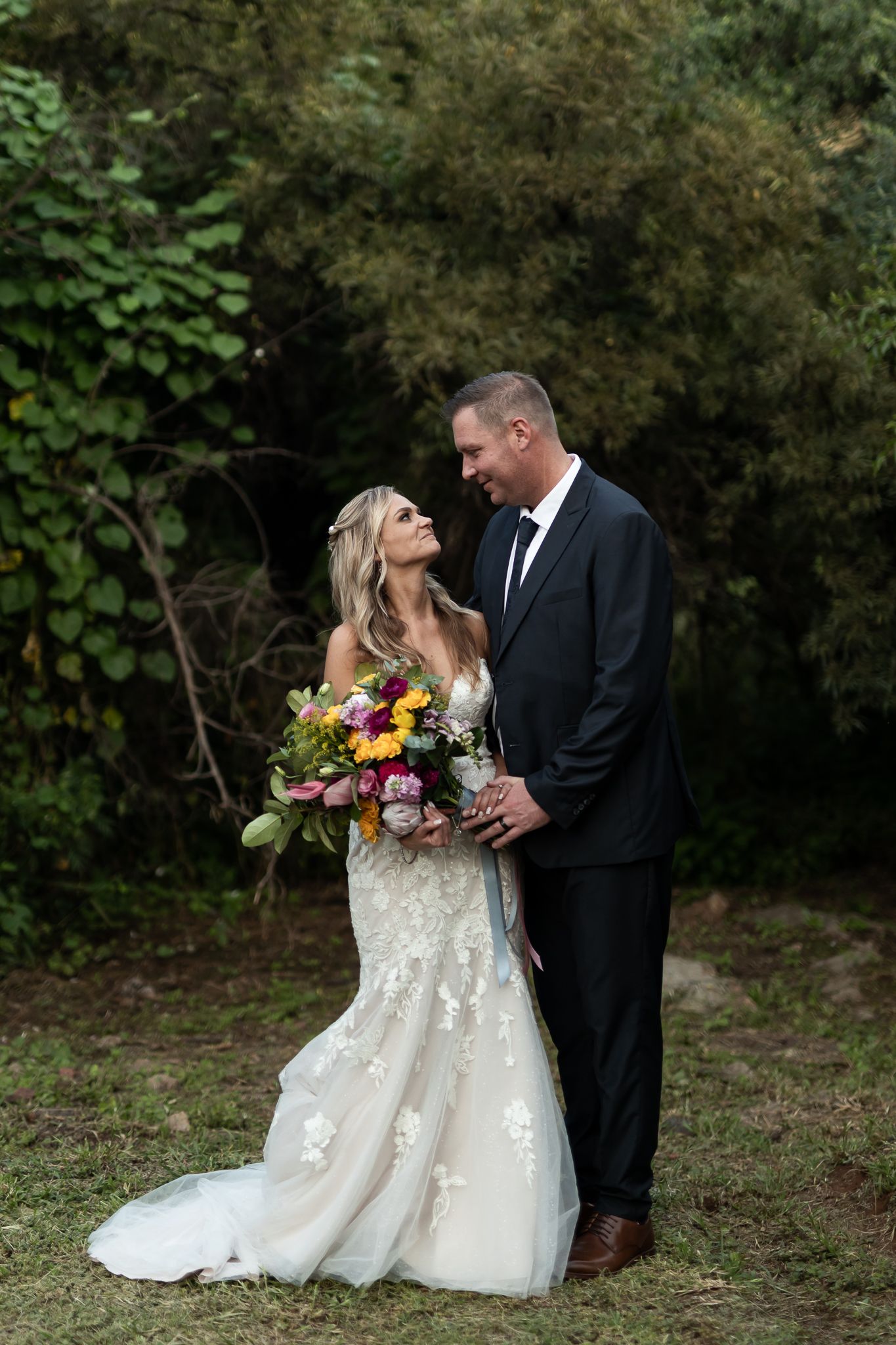 Wedding couple poses with vibrant wildflower bouquet against green foliage.