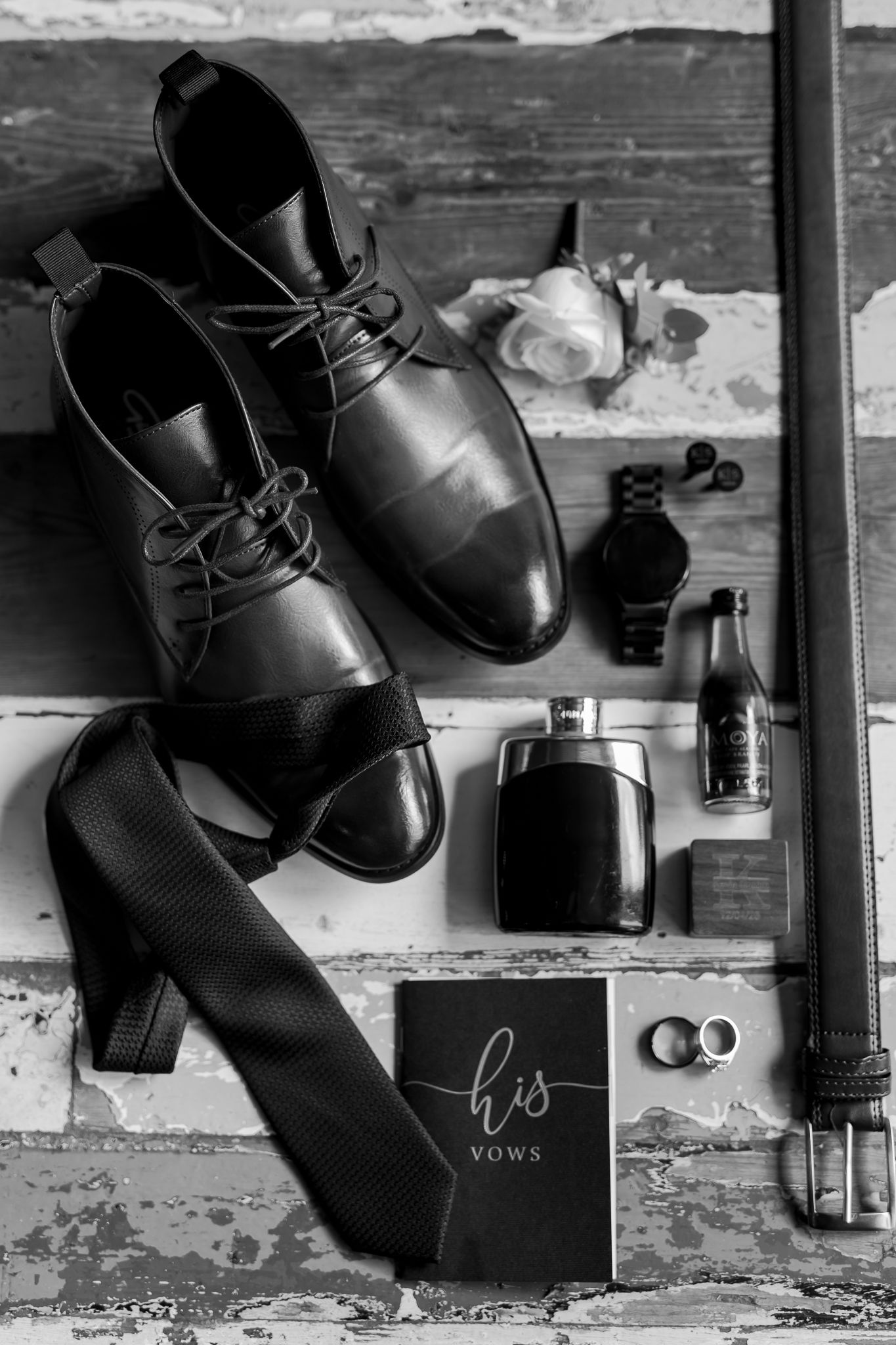 Wedding accessories including black dress shoes, tie, rings and cologne on rustic wooden surface.