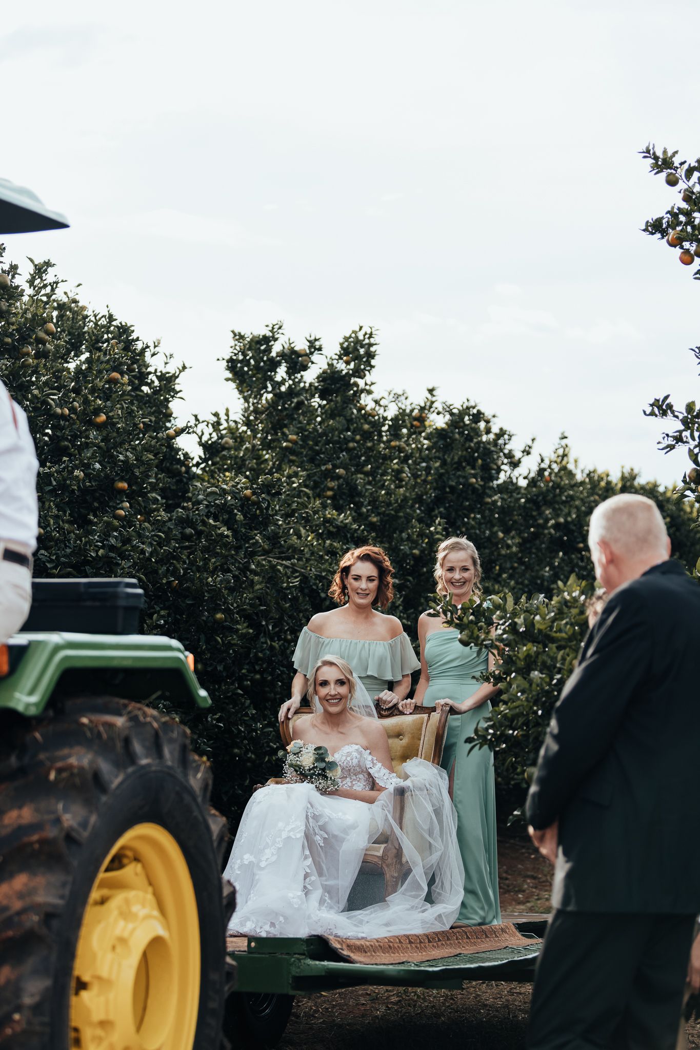 Wedding party poses on a vintage green tractor in an orchard setting during a rustic outdoor ceremony.