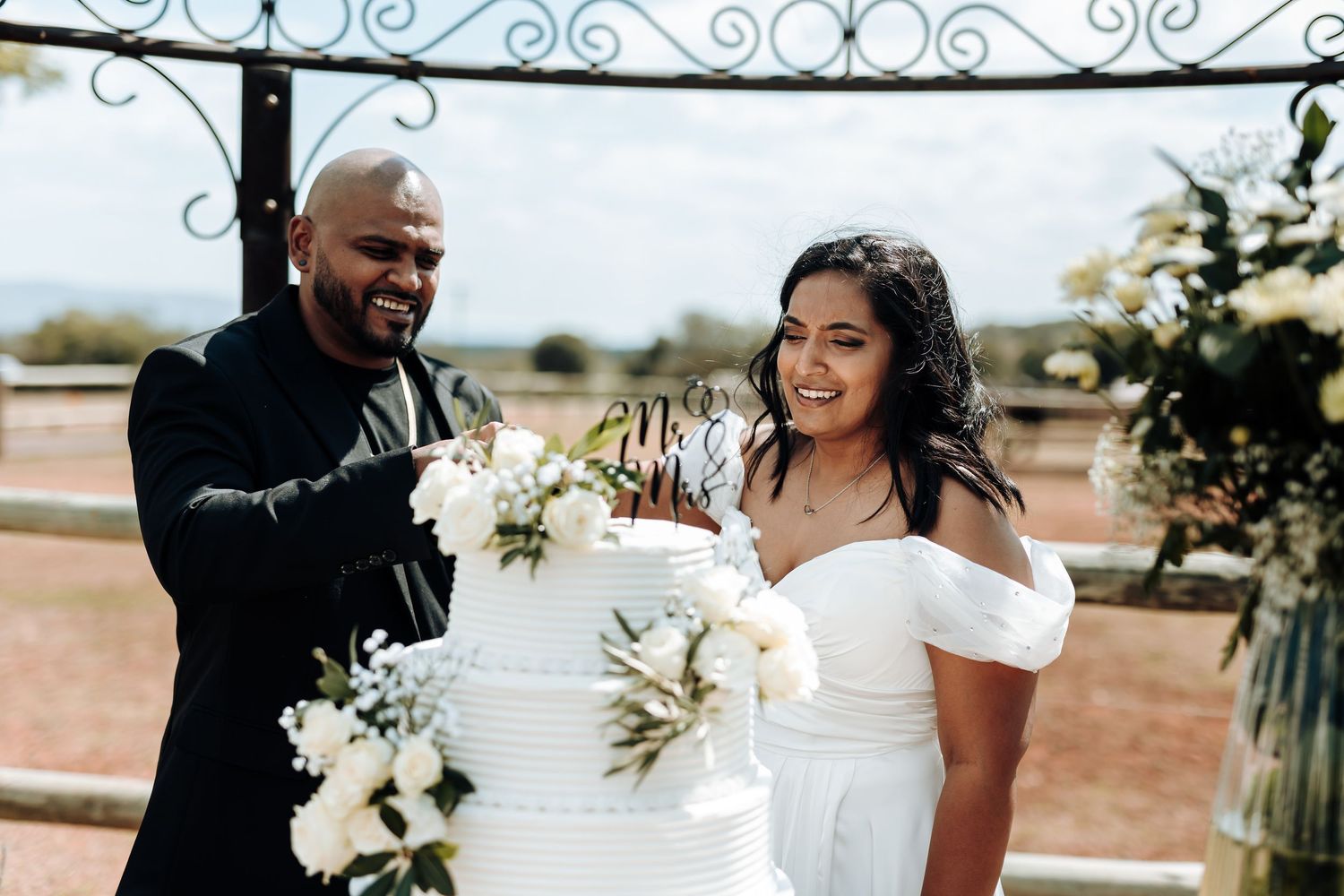 Happy couple cutting their white tiered wedding cake under decorative arch.