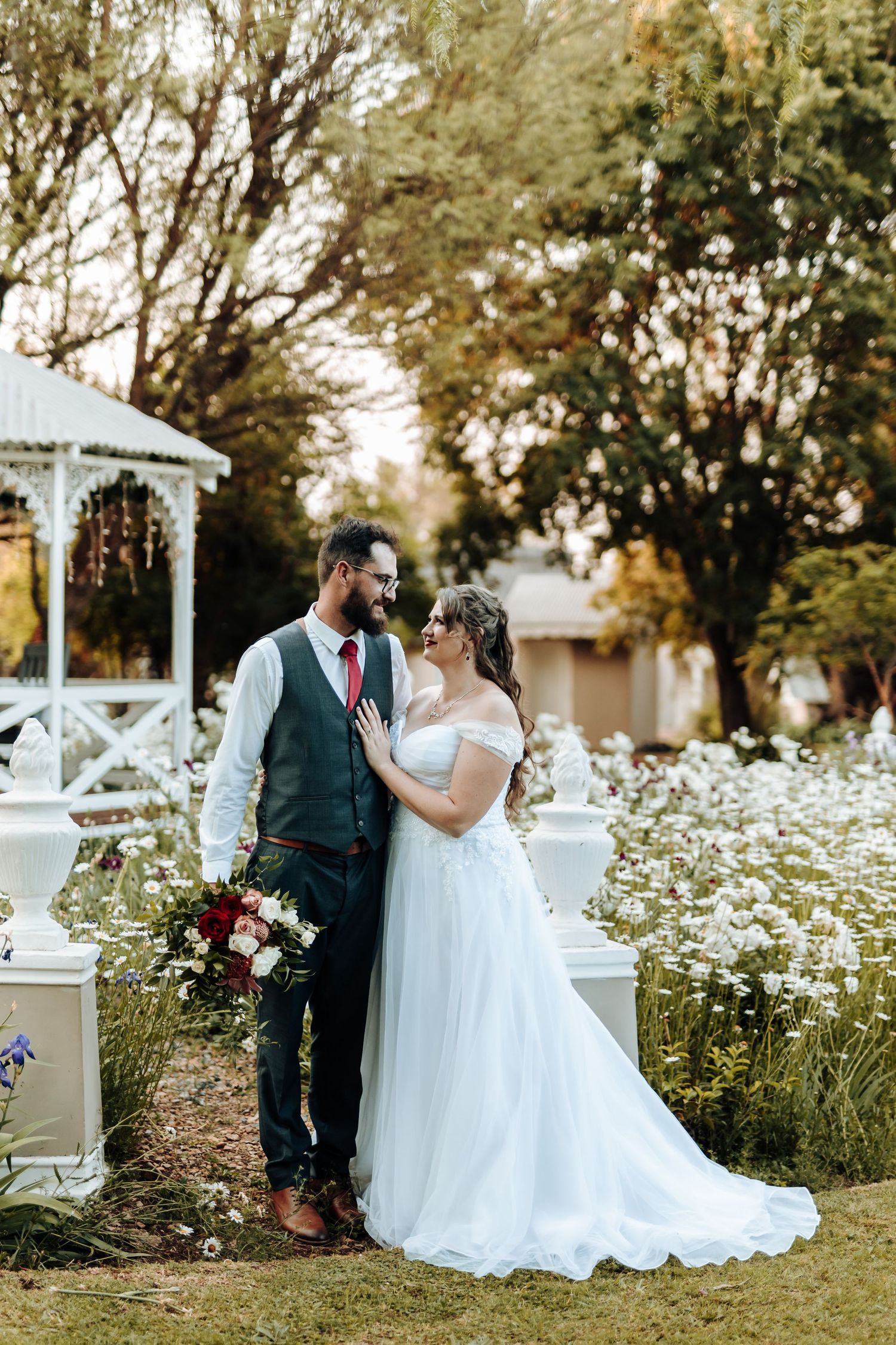 Wedding couple poses in garden setting with white gazebo and flowering meadow background.