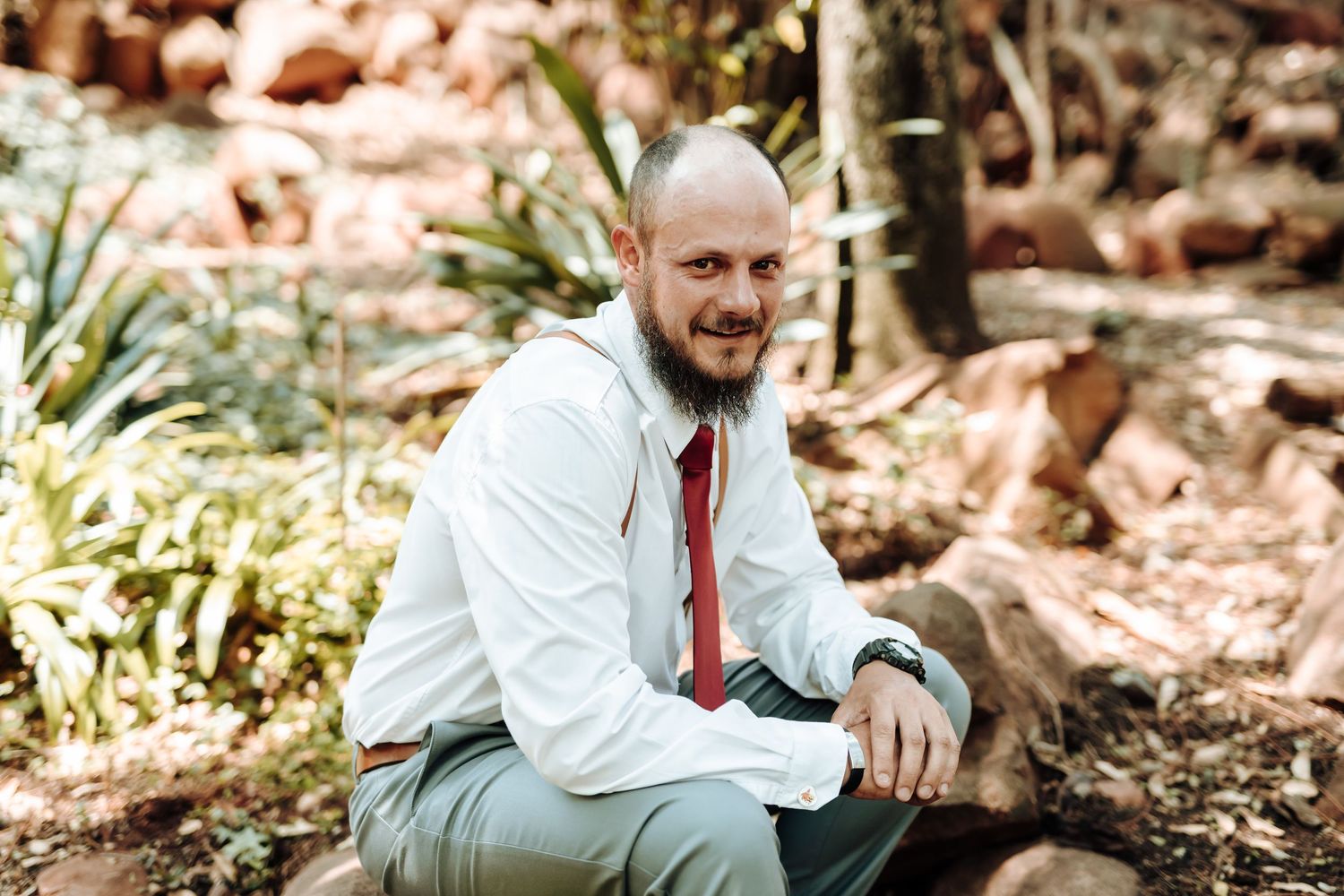 Single groomsman in white shirt and red tie sits for portrait in outdoor garden setting.