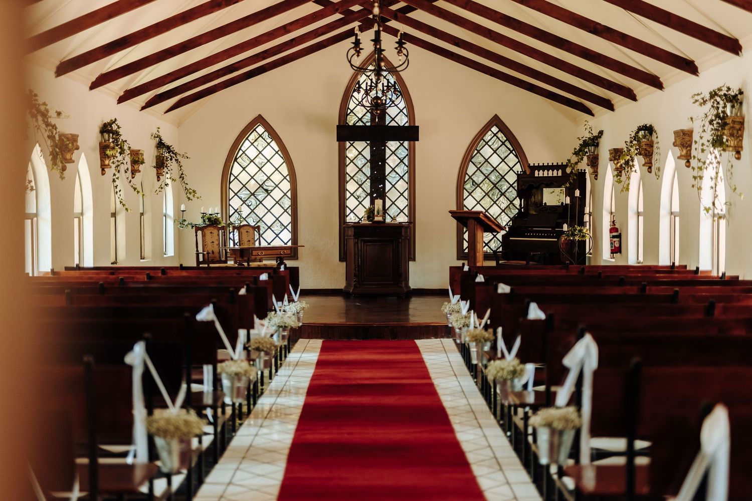 Church interior with gothic arched windows, wooden pews, and red carpet aisle runner.