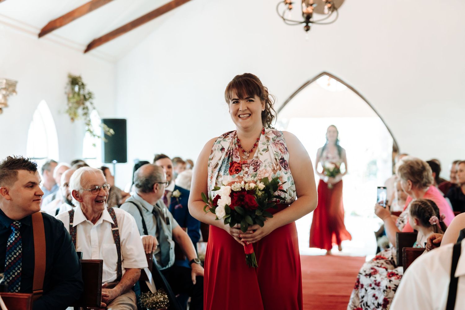 Bridesmaid in red dress holding bouquet during indoor wedding ceremony.