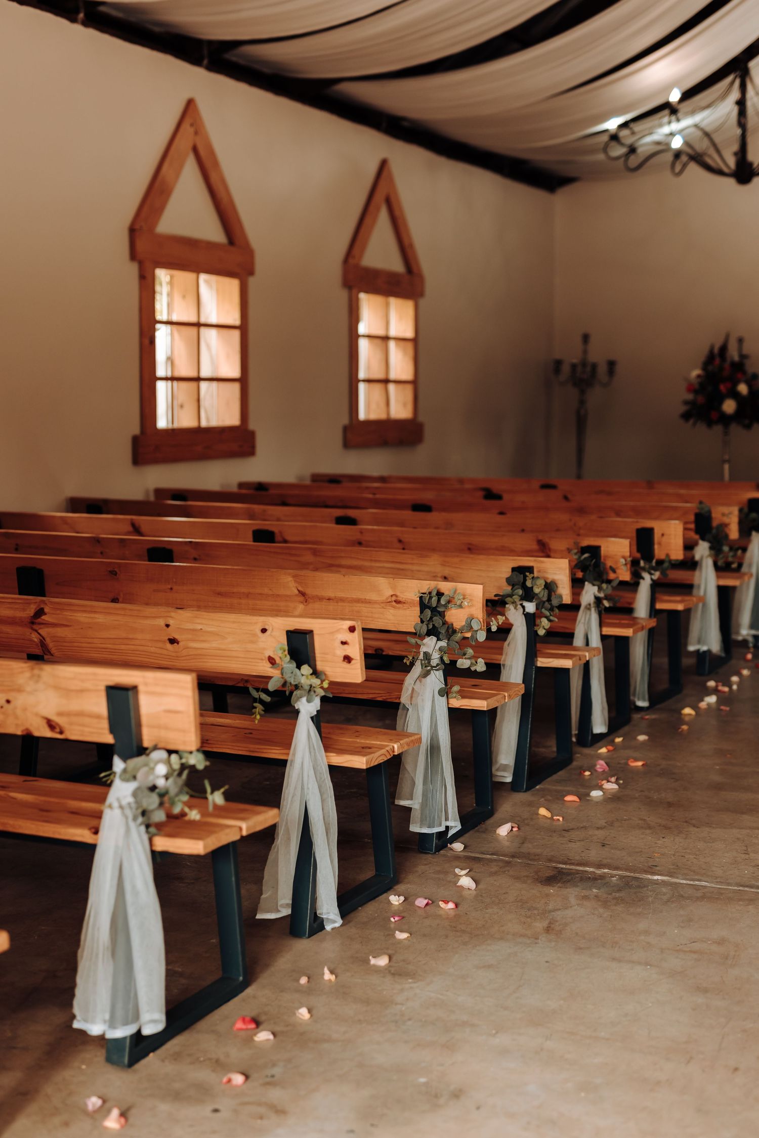 Church pews decorated with white fabric bows and glowing candles line the aisle of a charming chapel.