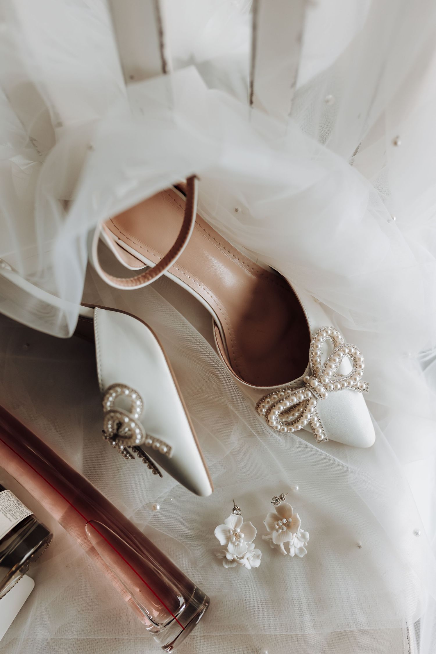 White bridal shoes with pearl bow embellishments and earrings on a delicate tulle background.