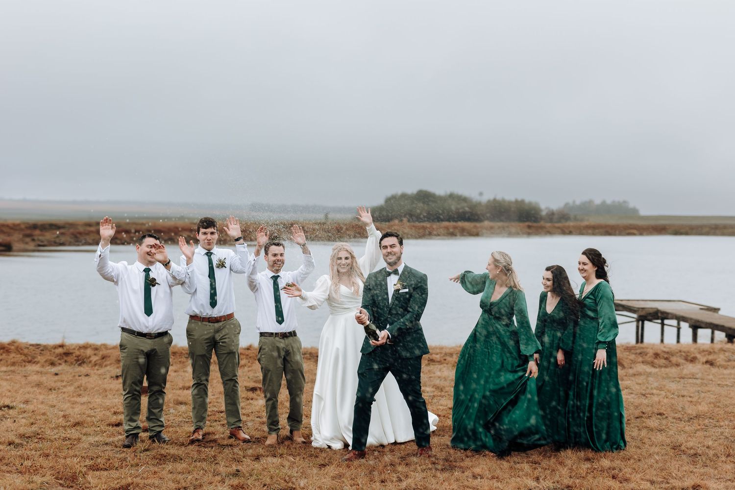 Wedding party cheers and celebrates together on a wooden dock by the water.