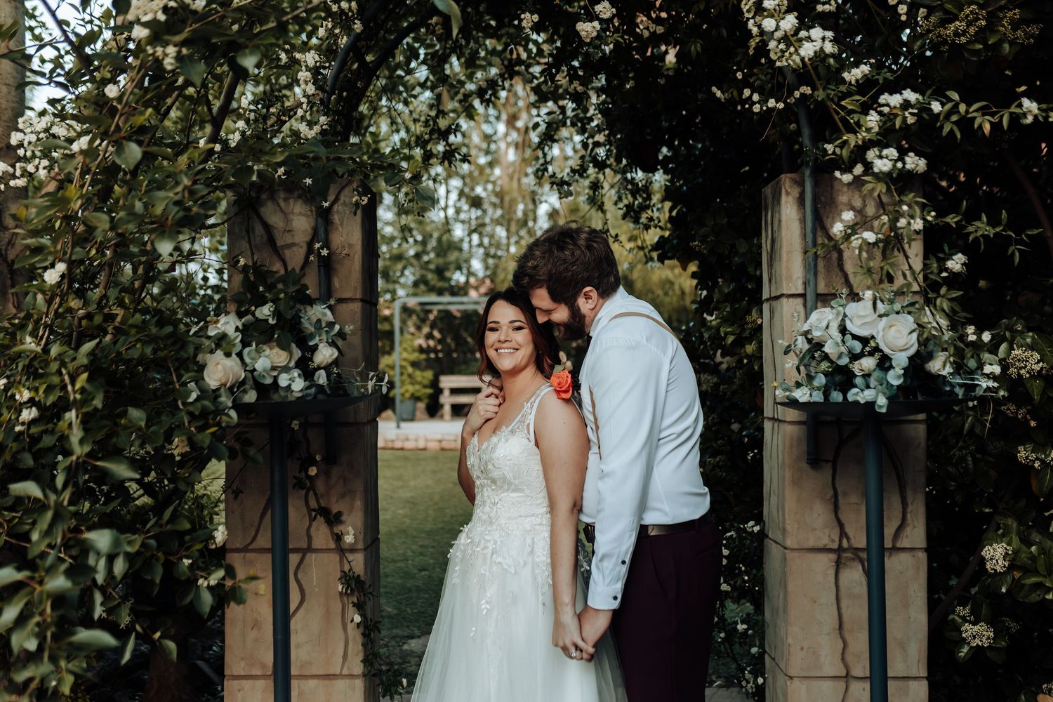 Couple holding hands under decorated garden archway during outdoor ceremony.