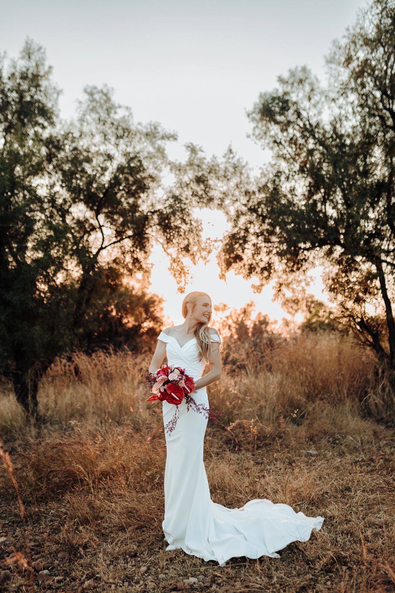 A bride in a flowing white gown stands in golden light holding red flowers.