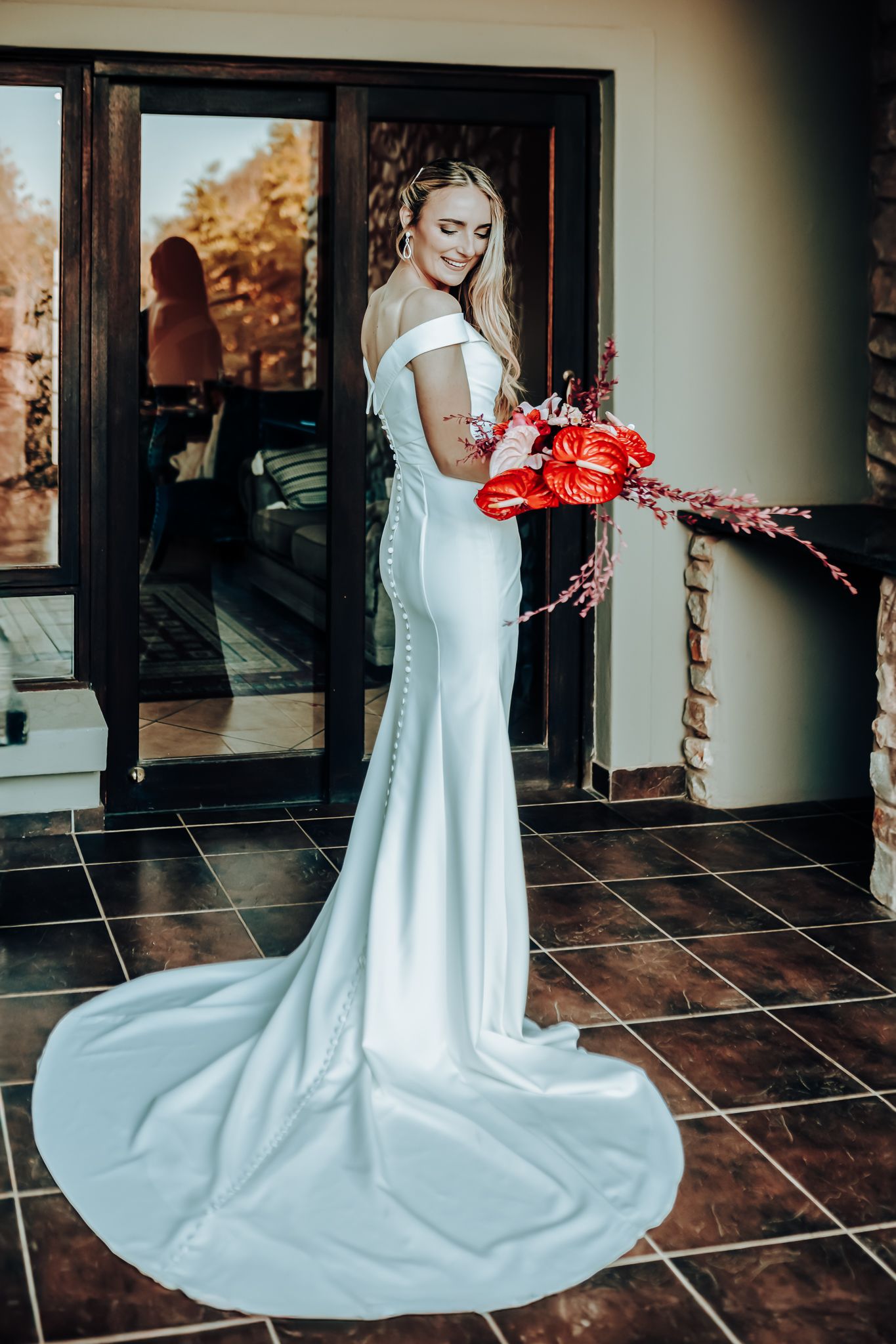 Bride in form-fitting white gown with dramatic train holding red floral arrangement.