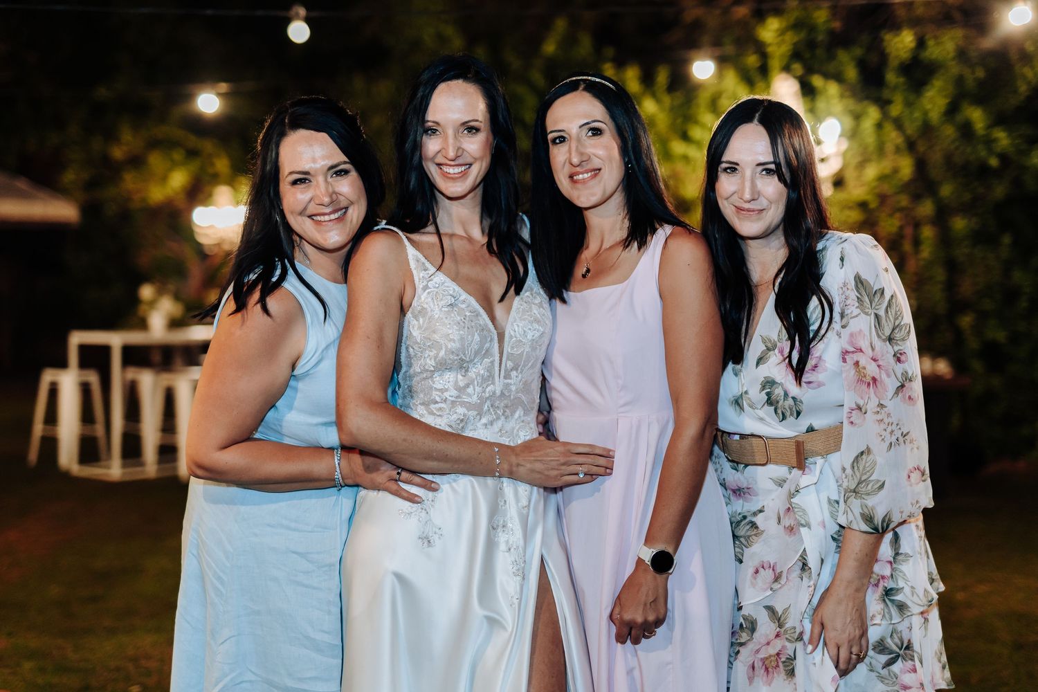 Four women in elegant dresses smiling together at an outdoor evening wedding celebration.