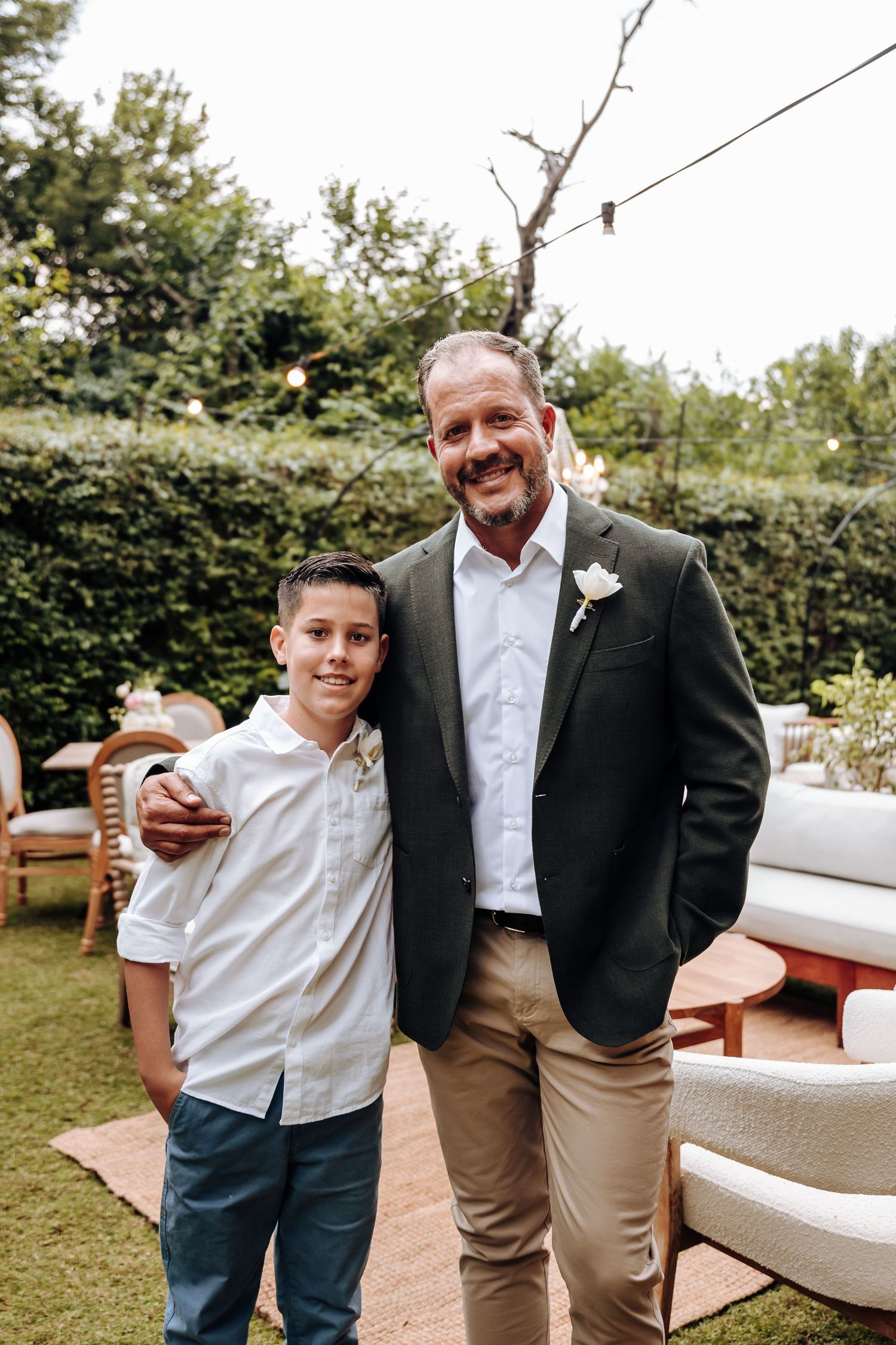 A man in a dark blazer with a boutonniere poses with a young boy in a white shirt at an outdoor event.